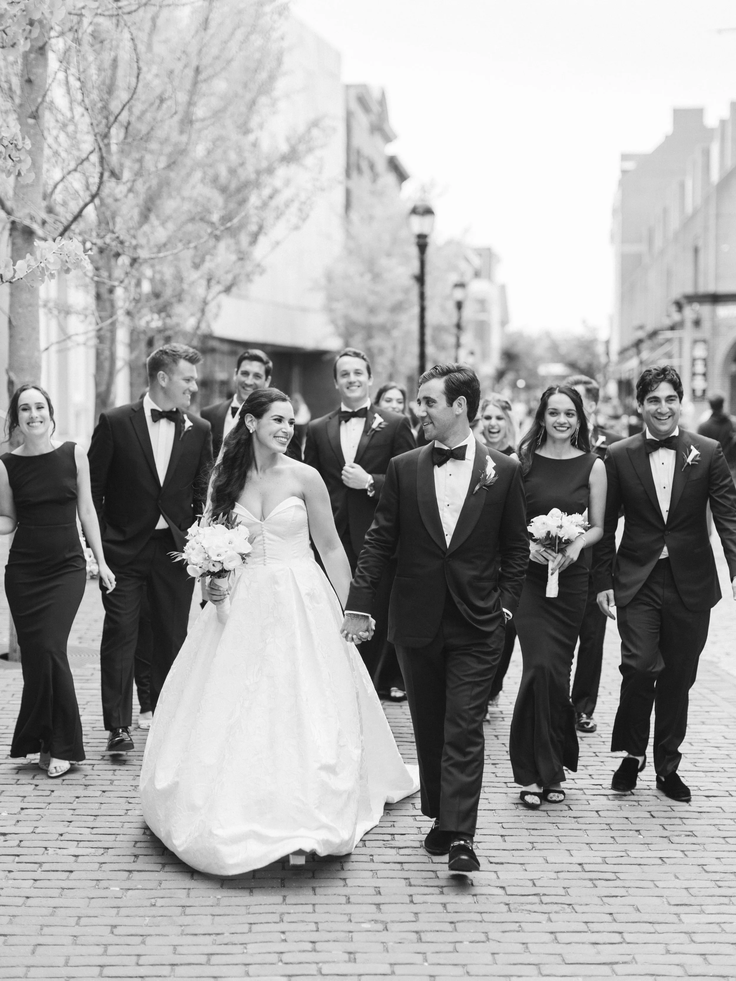 Black and white photo of a bride and groom walking hand in hand in front of a group of friends during a wedding celebration on a city street, smiling and dressed formally.