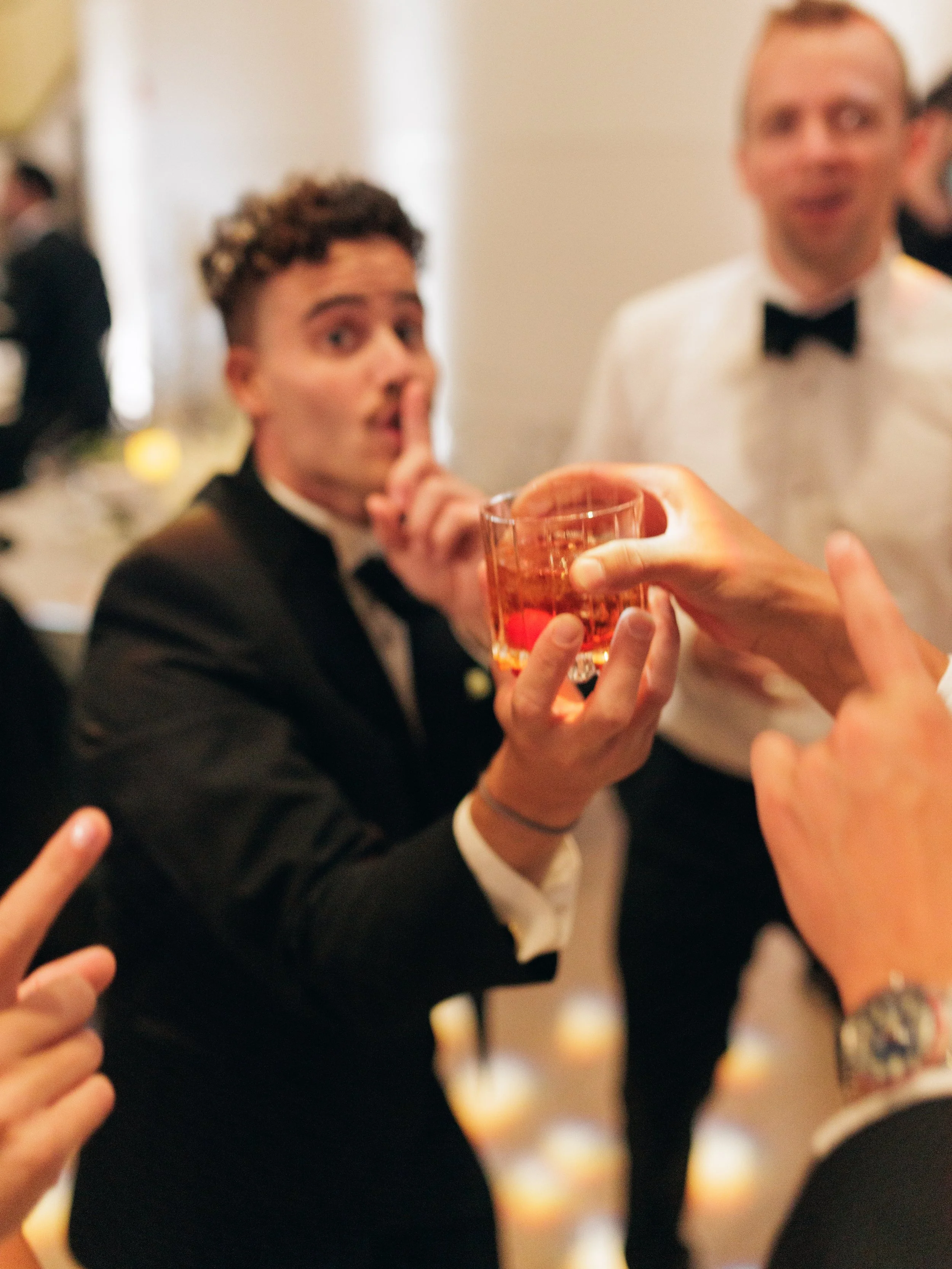 People at a formal event with a woman in a tuxedo and a man in a tuxedo, someone is offering a drink in a glass.
