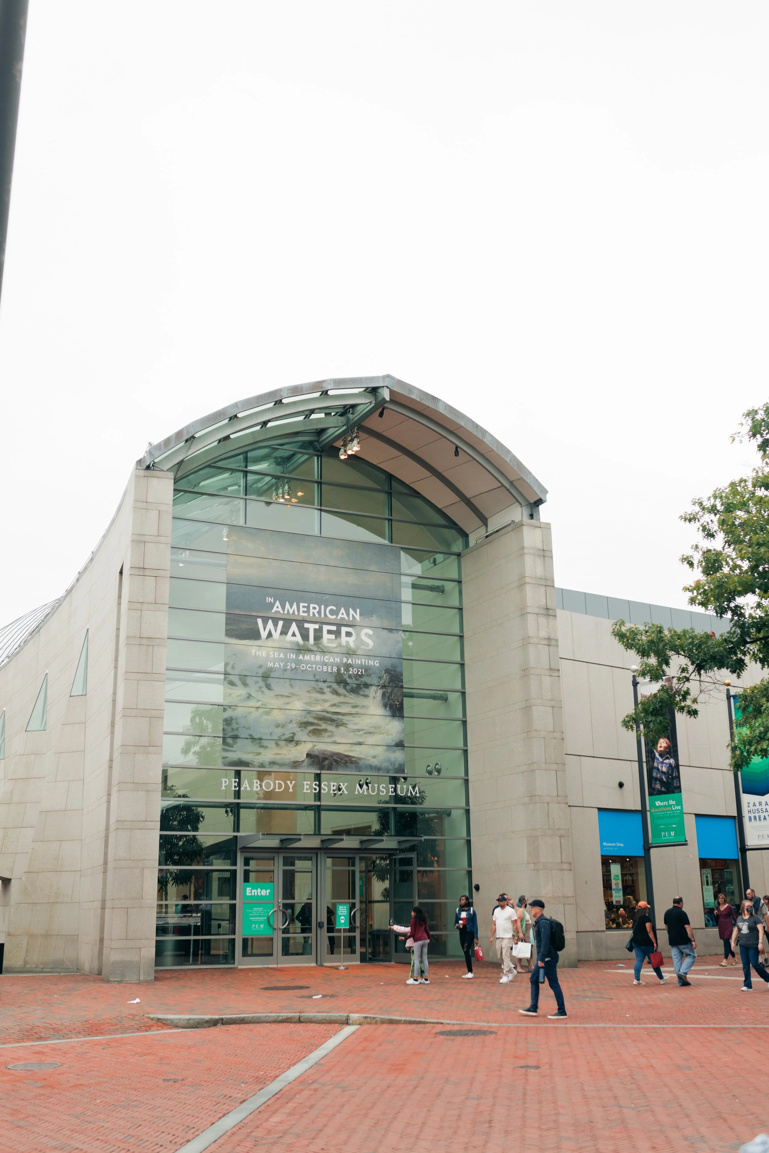 People walking in front of the Peabody Essex Museum entrance with a banner advertising the 'In American Waters' exhibition.