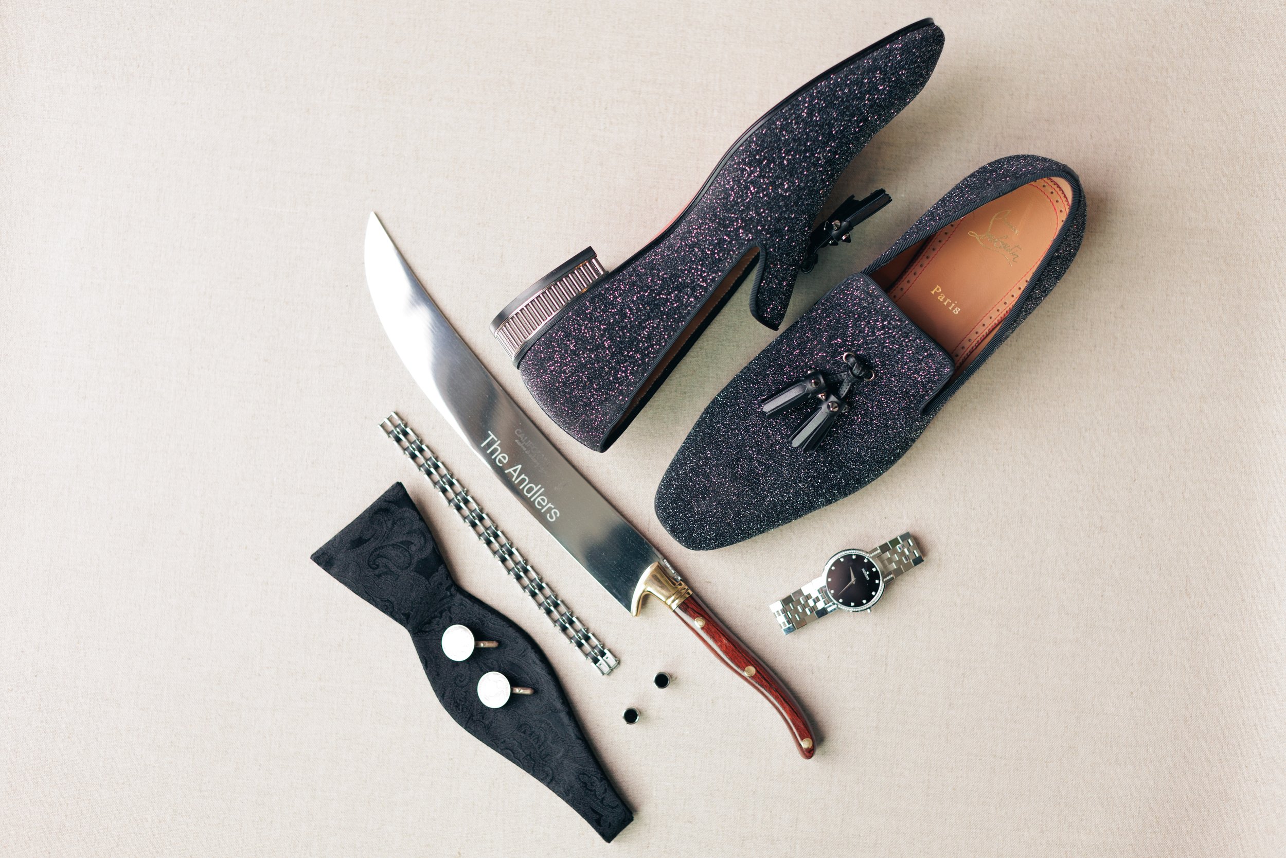 Black glittery high heel shoe with a zipper on the back, placed on a beige surface along with jewelry, a knife, and a book.