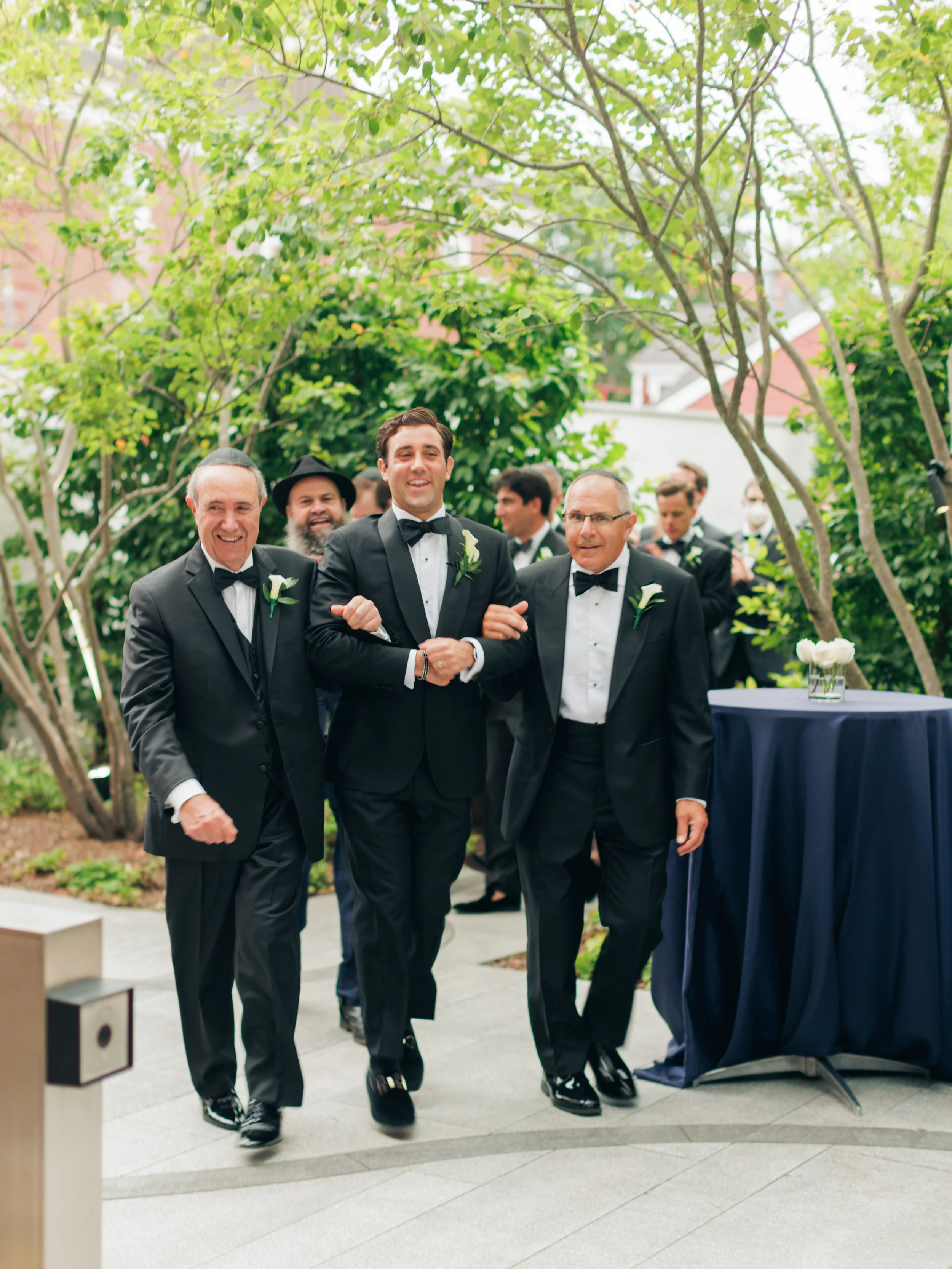 Group of men dressed in black tuxedos and bow ties, walking together outdoors under green trees, at a formal event or wedding.