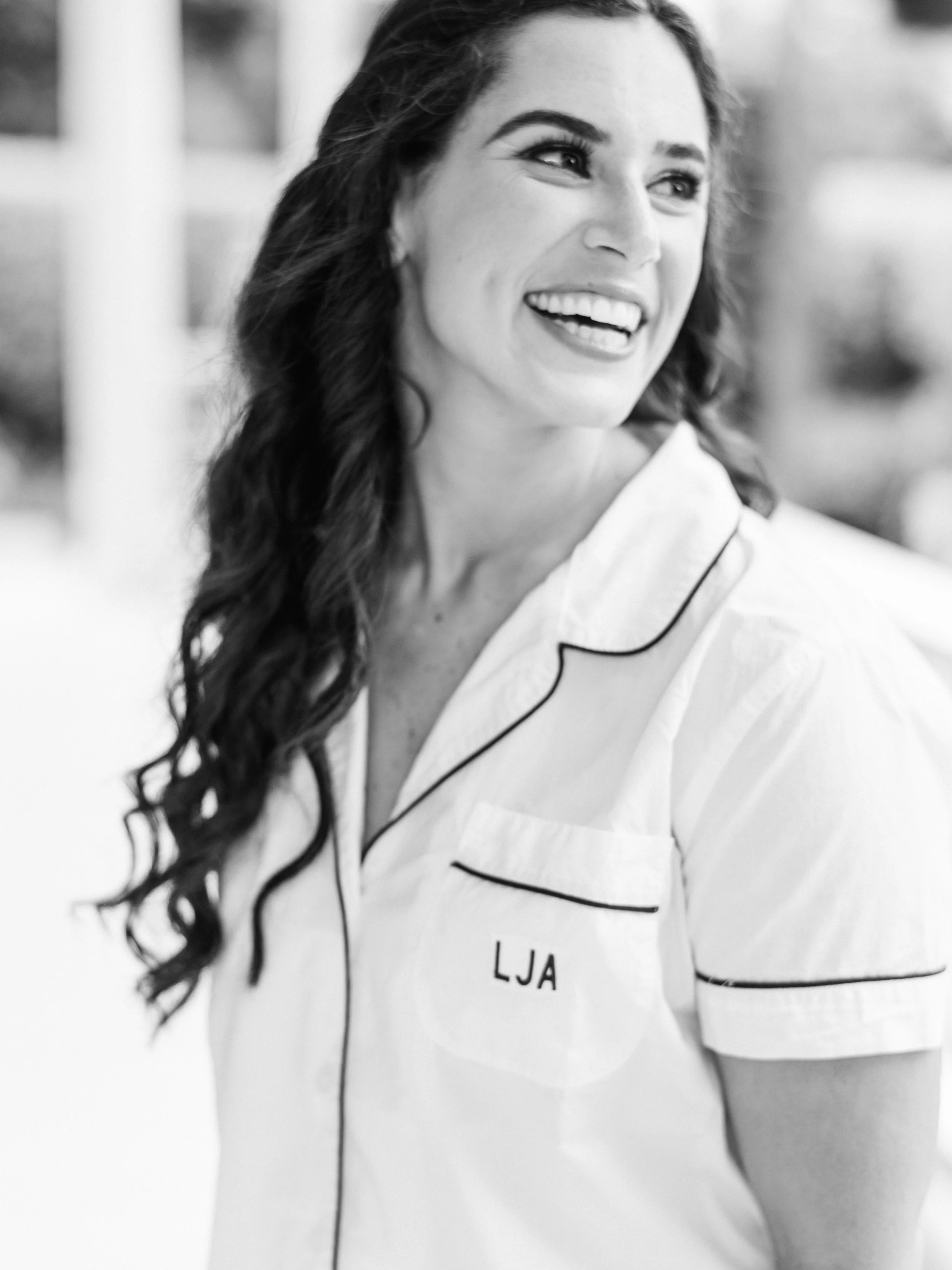 Woman with long dark hair, wearing a jacket with the initials 'LJA' on it, smiling and looking to her left, in black and white.