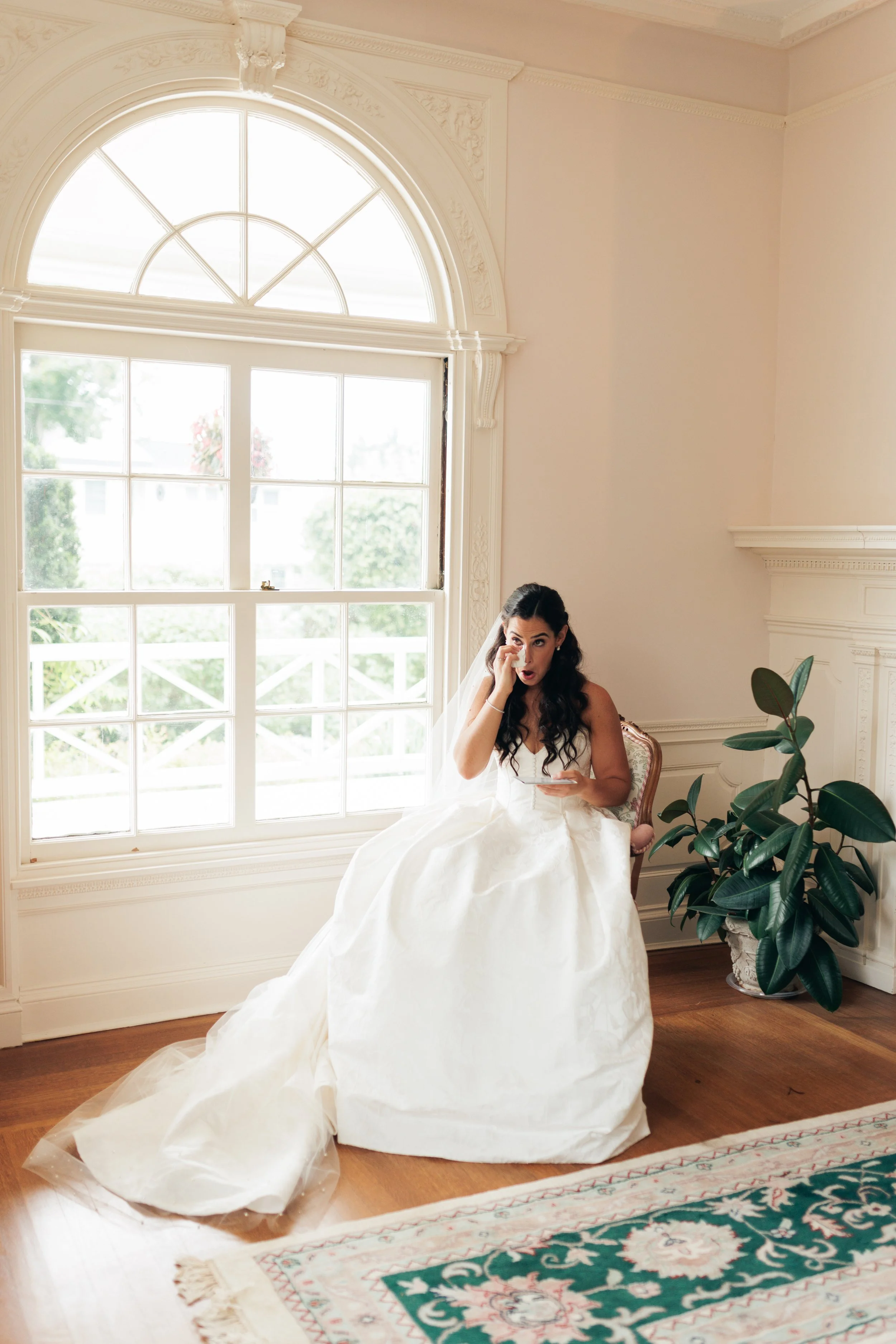 A woman in a wedding dress sitting on a chair in a room with large window, decorative molding, and a potted plant.