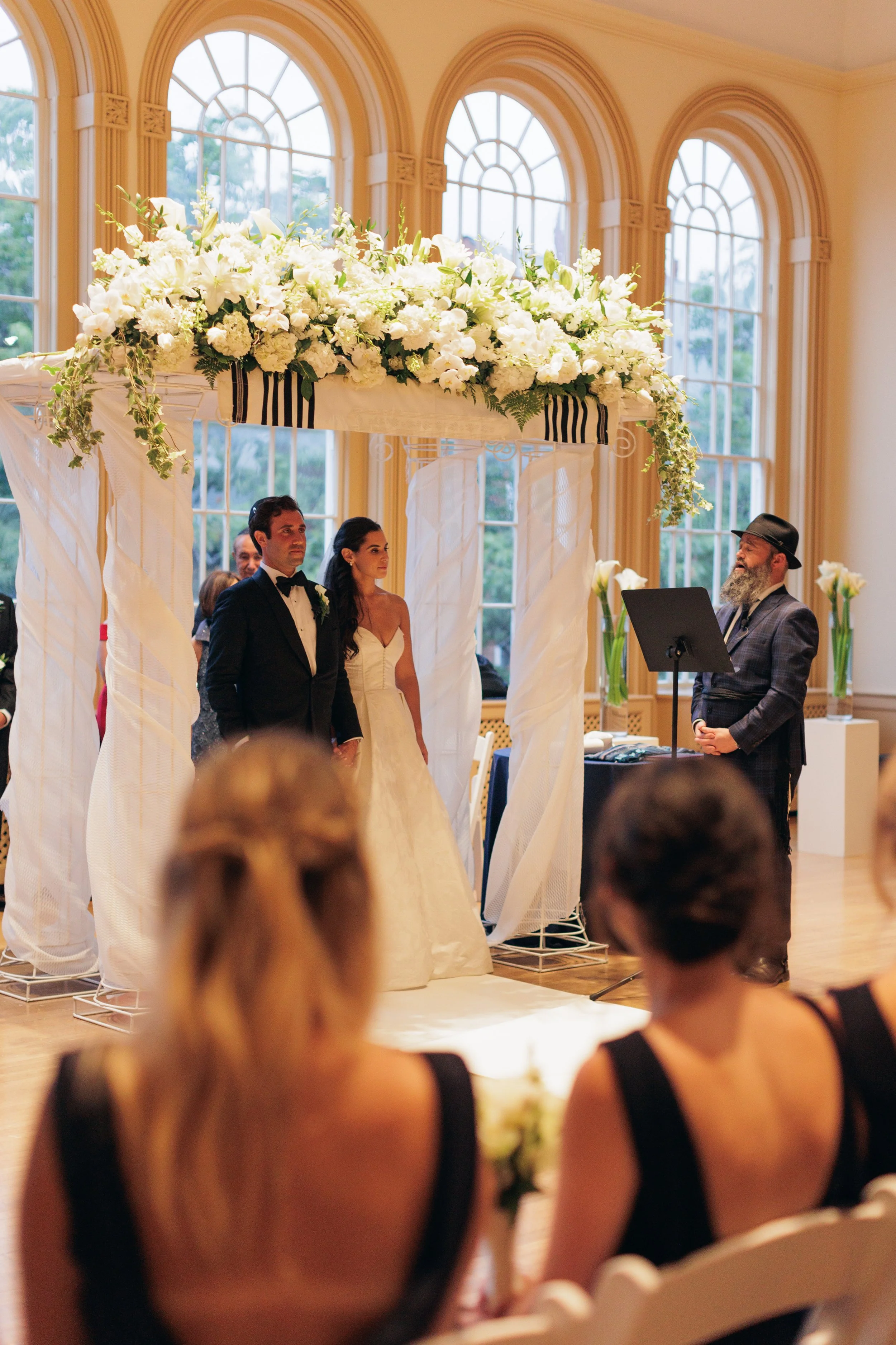 A wedding ceremony in a large, elegant room with high arched windows. The couple, dressed in wedding attire, stands under a floral arch. A man with a beard and fedora stands nearby, possibly officiating. Guests are seated in the foreground.