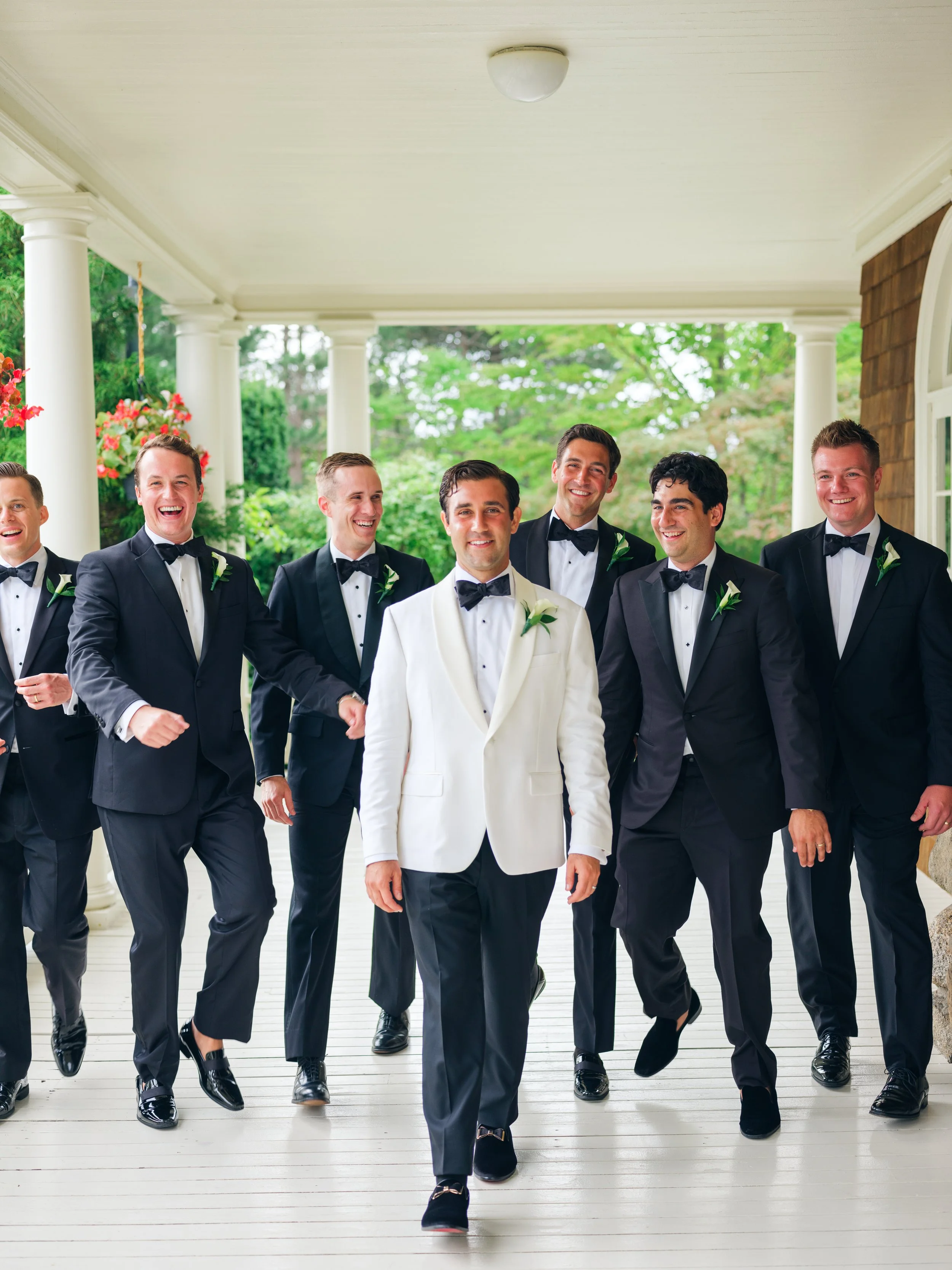 A group of men dressed in tuxedos, walking confidently on a porch with white pillars and greenery in the background.