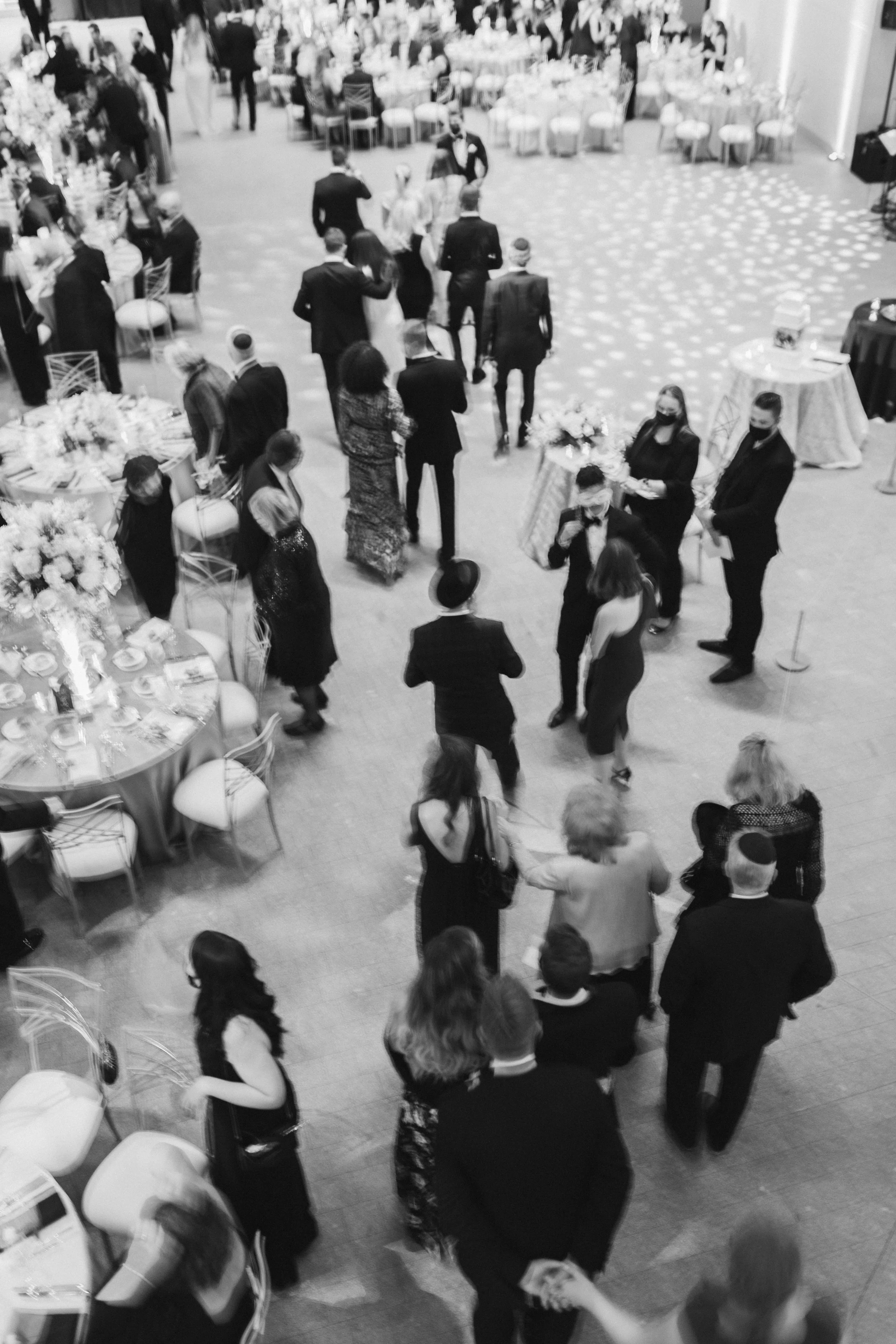 Guests dancing and socializing at a formal event or wedding reception in a large banquet hall with decorated tables and floral arrangements.