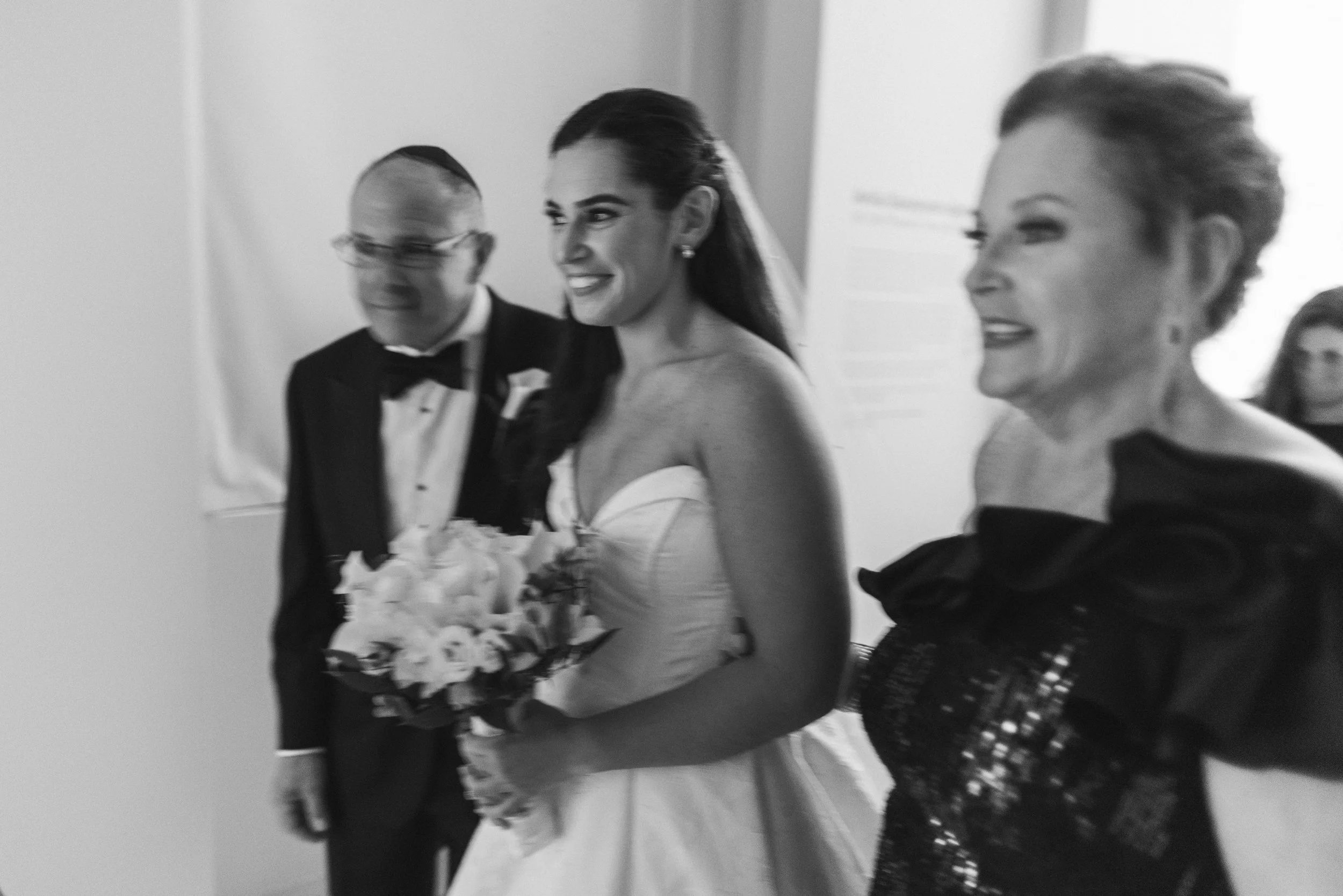 A bride in a strapless wedding gown holding a bouquet, walking down the aisle with a wide smile, accompanied by two women and a man dressed in formal attire, in a wedding ceremony.