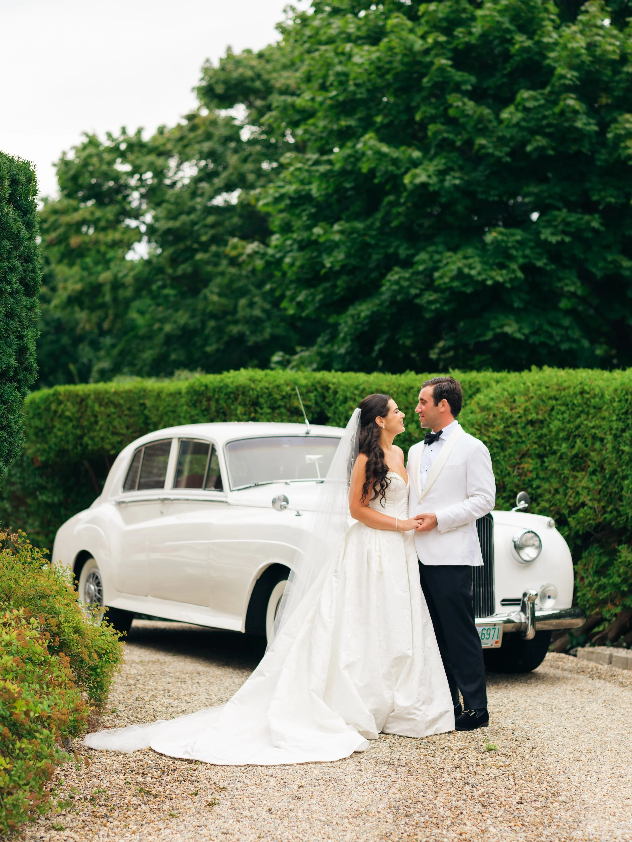 A bride and groom standing close together near a white vintage car, surrounded by greenery, during their wedding celebration.
