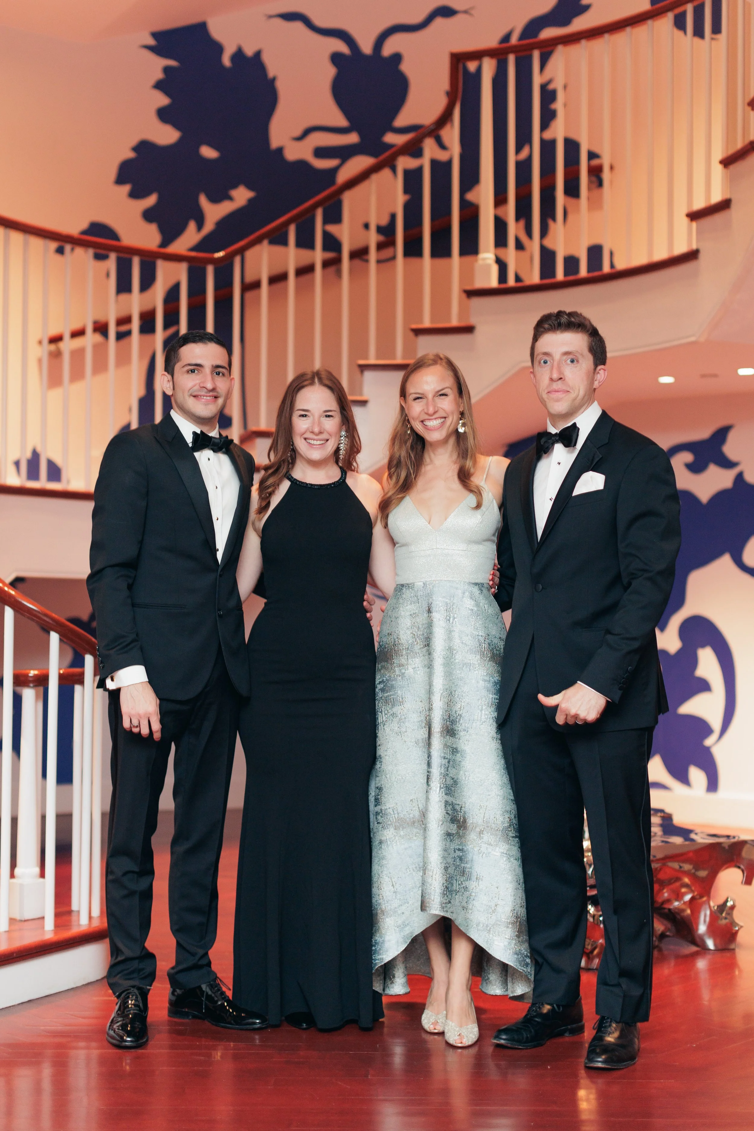 Four people dressed in formal attire standing on a staircase at a fancy event. The two men are wearing tuxedos with bow ties, and the two women are wearing elegant dresses. They are smiling and looking at the camera. The background features a large w