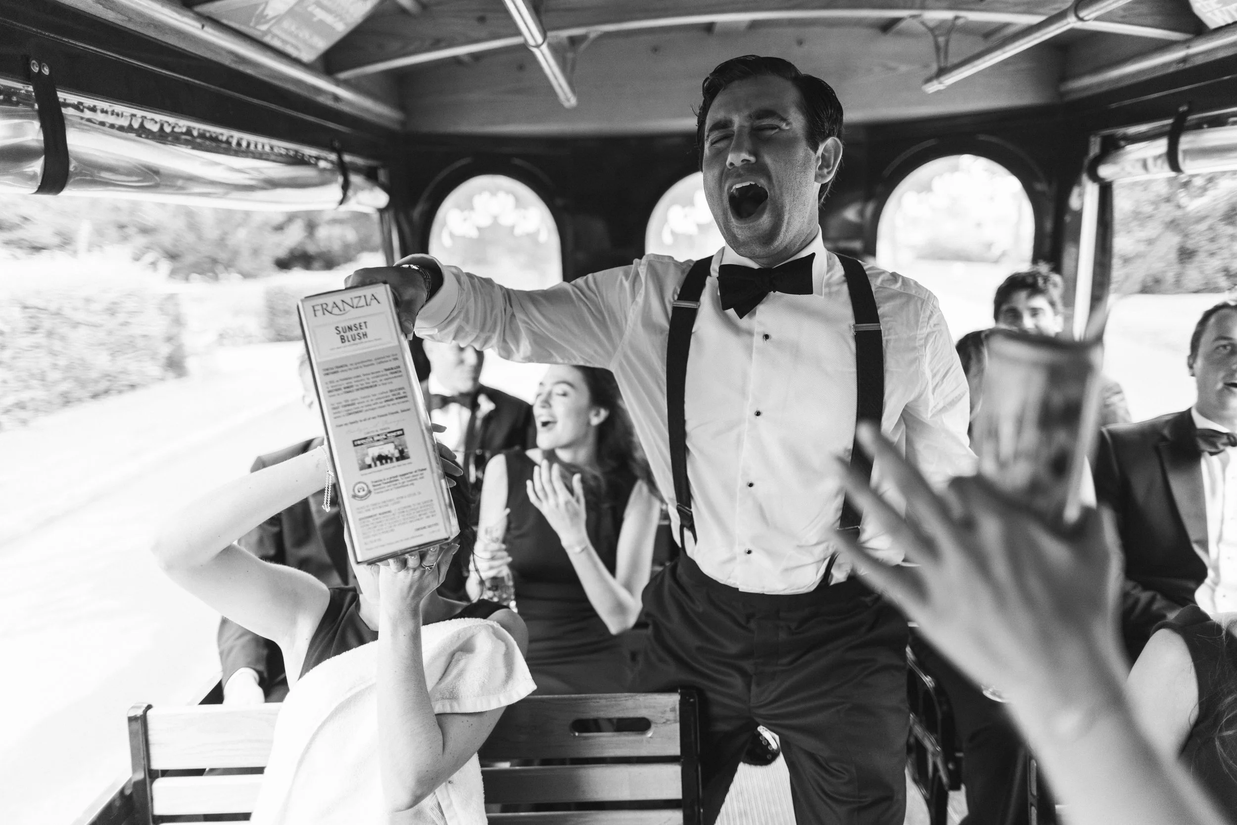 A man dressed in a tuxedo with suspenders and a bowtie, smiling and singing loudly on a trolley with a group of people, some sitting and some standing, during a celebration or social gathering.