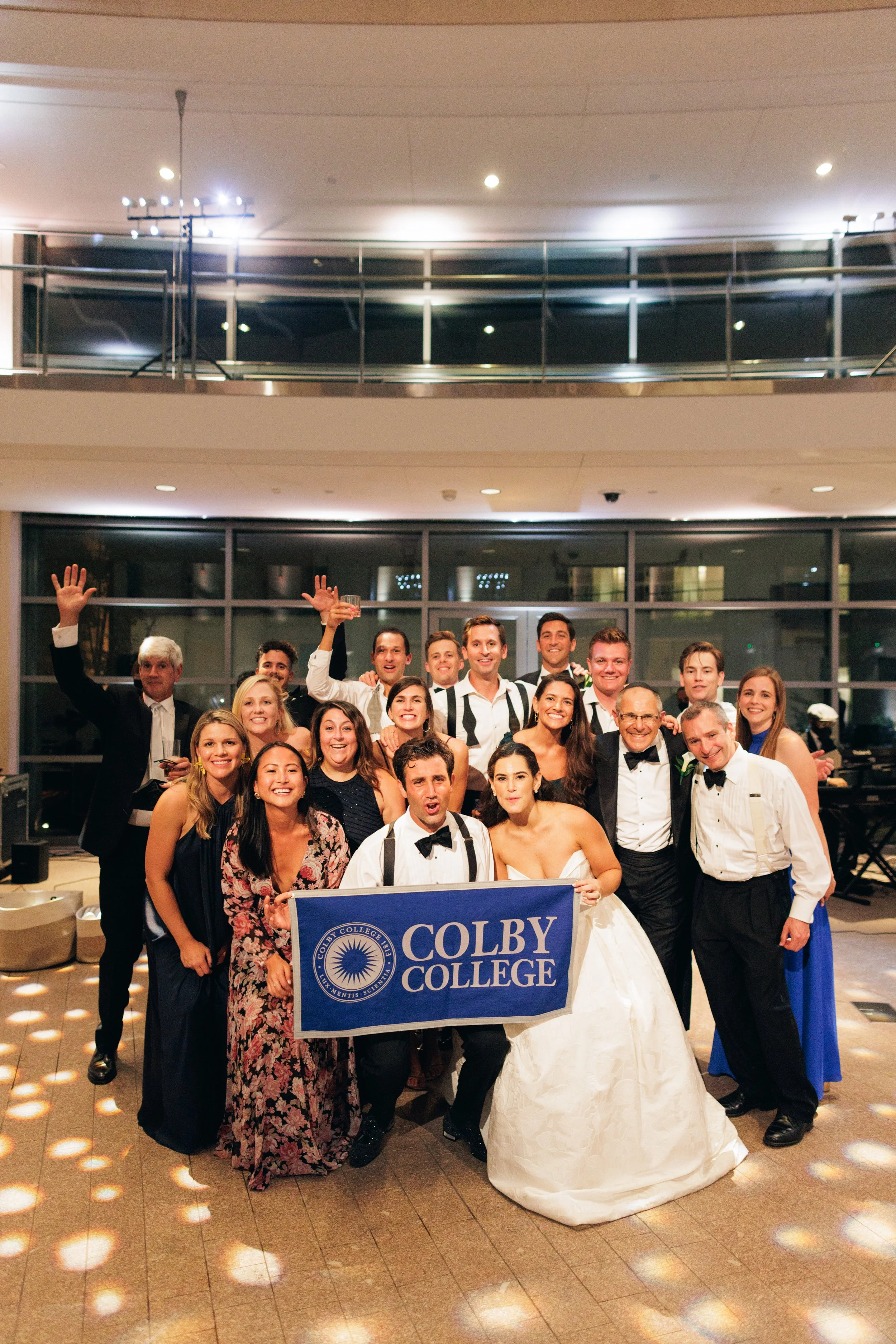Group of people at a graduation celebration holding a Colby College banner inside a ballroom, with a bride in a wedding dress in front.