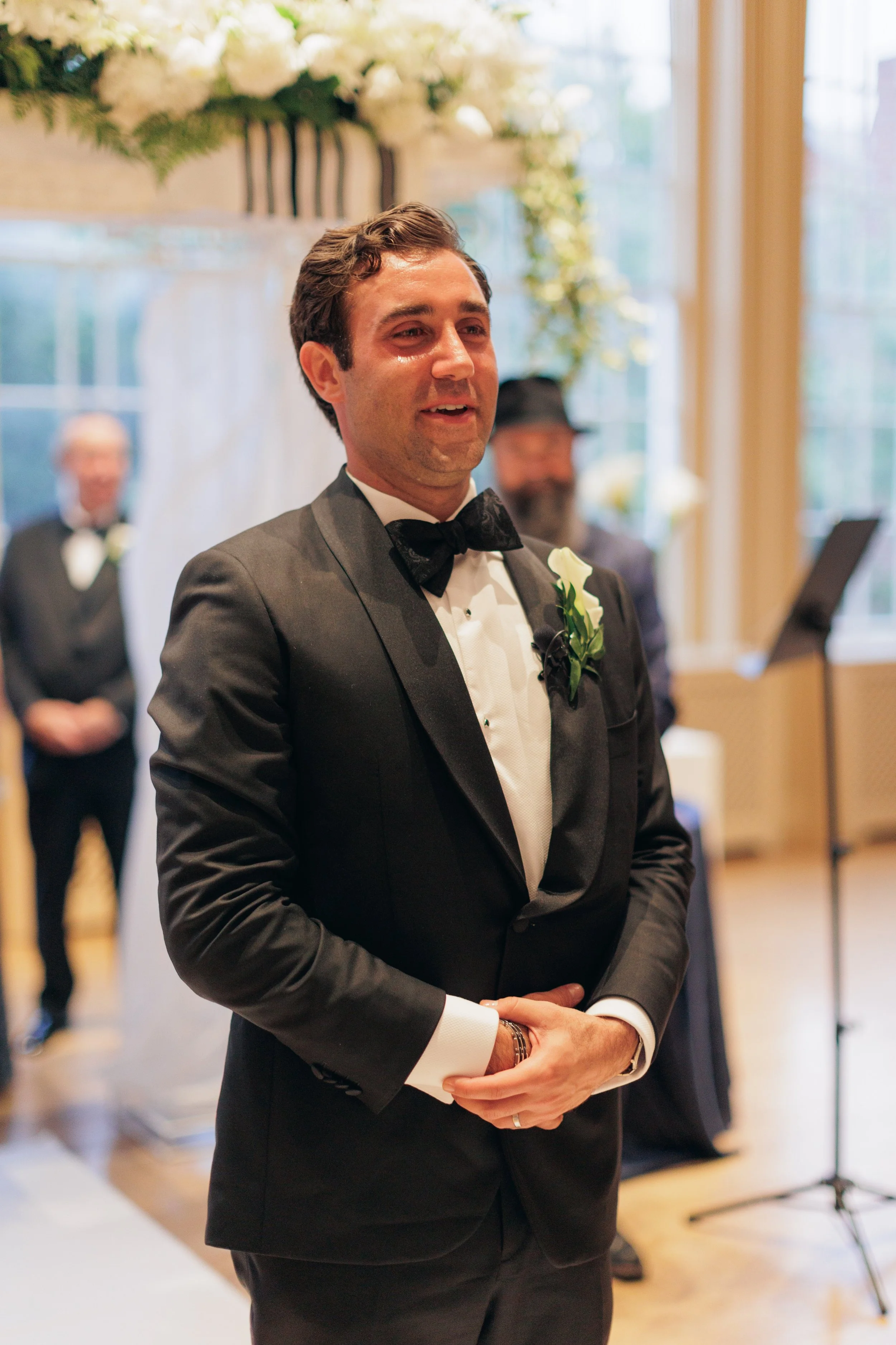 A man in a black tuxedo with a bow tie and boutonniere, standing indoors during a wedding, with guests and floral arrangements in the background.