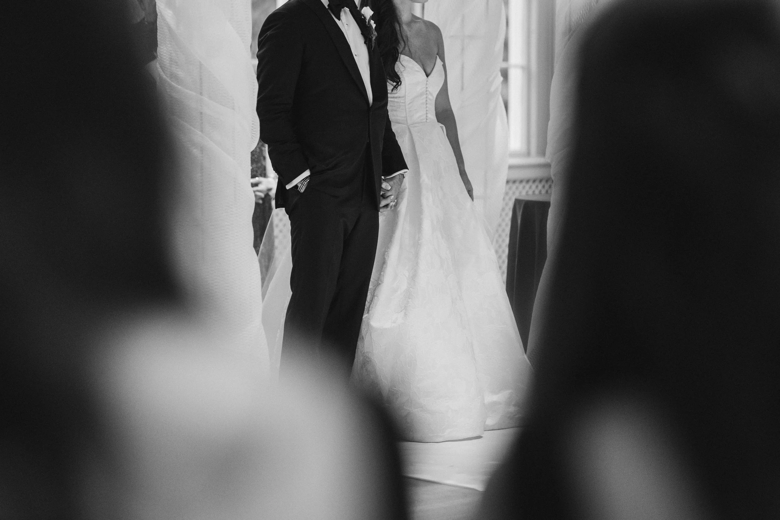 A wedding ceremony scene, with a bride in a white gown and a groom in a tuxedo, standing together in a room with large windows, seen through a partially blocked camera angle.