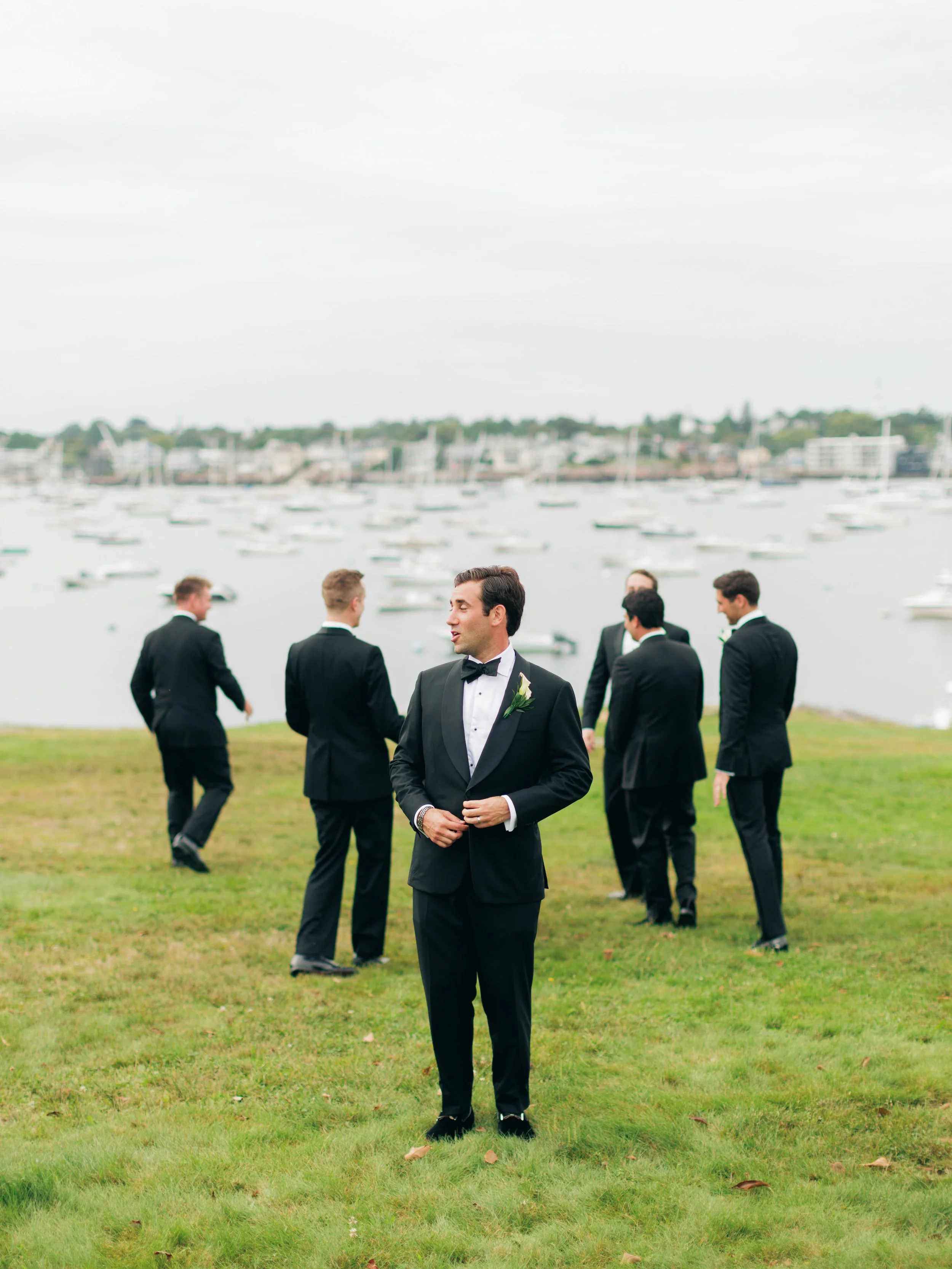 Group of men in tuxedos standing on grass near water with boats and buildings in background.