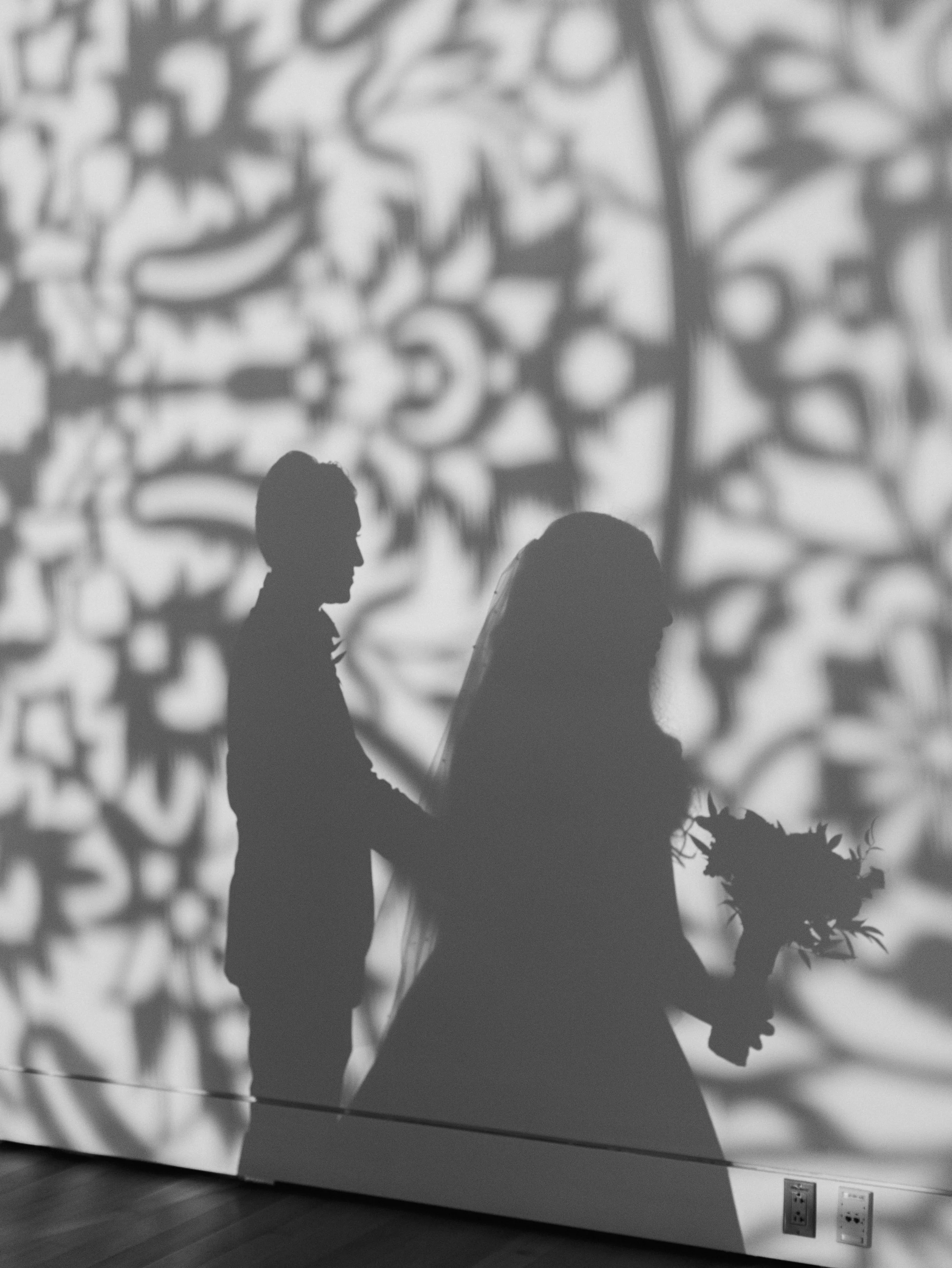 Silhouettes of a man and a woman holding hands, standing in front of a decorative wall with intricate patterns, in black and white.