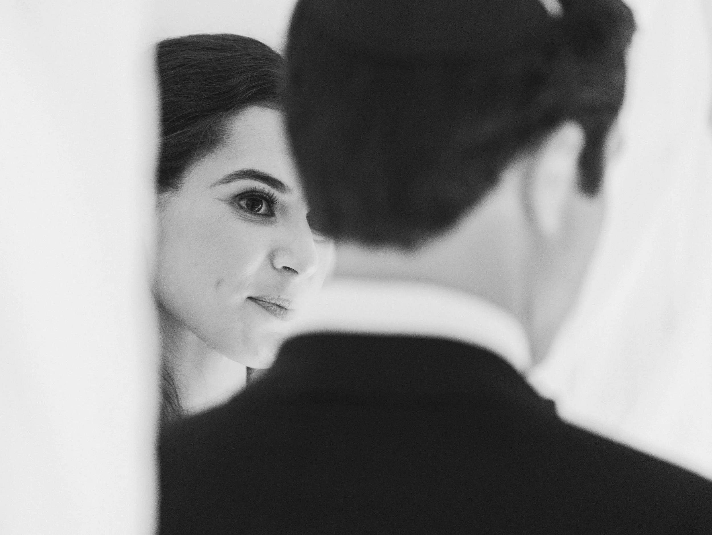 A woman with dark hair and a man in black clothing or cap face each other, with the woman's face partially visible, looking at each other. The image is in black and white.