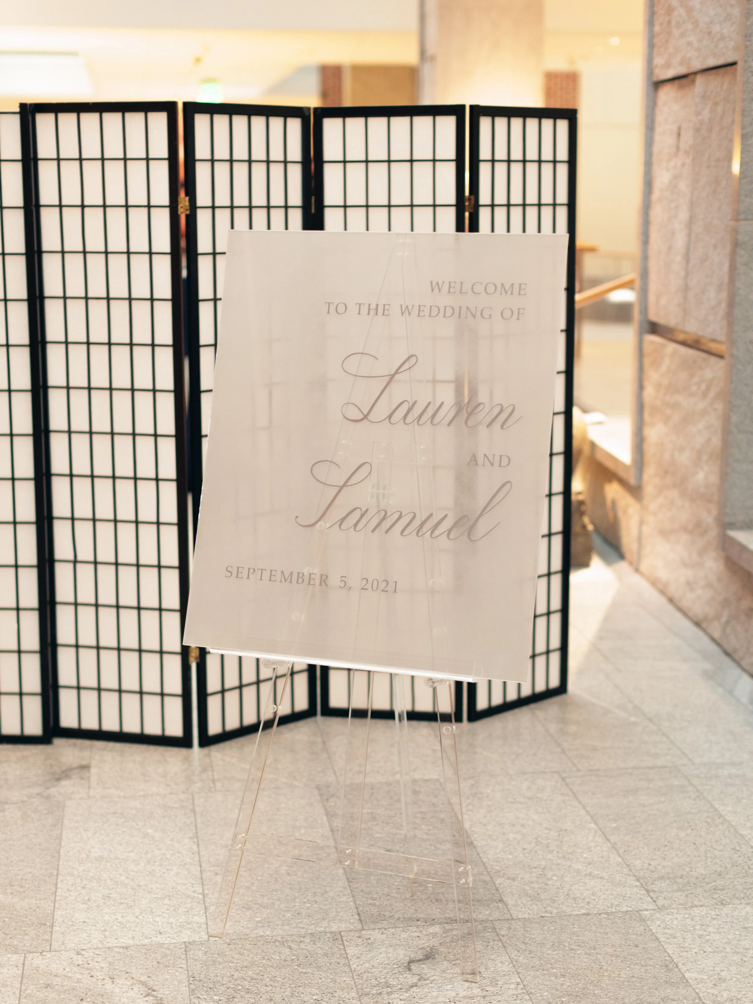 A decorative stand with a welcome sign for the wedding of Lauren and Samuel on September 5, 2021, placed in front of a black folding screen indoors.