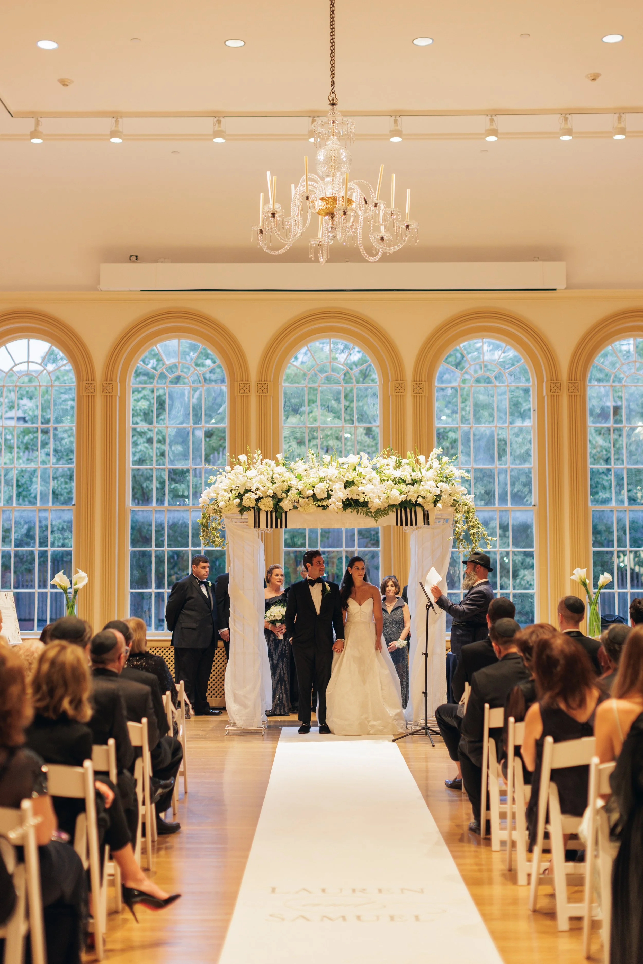 A diverse group of people getting married in a large, elegant room with tall windows and a chandelier, with guests seated on either side.