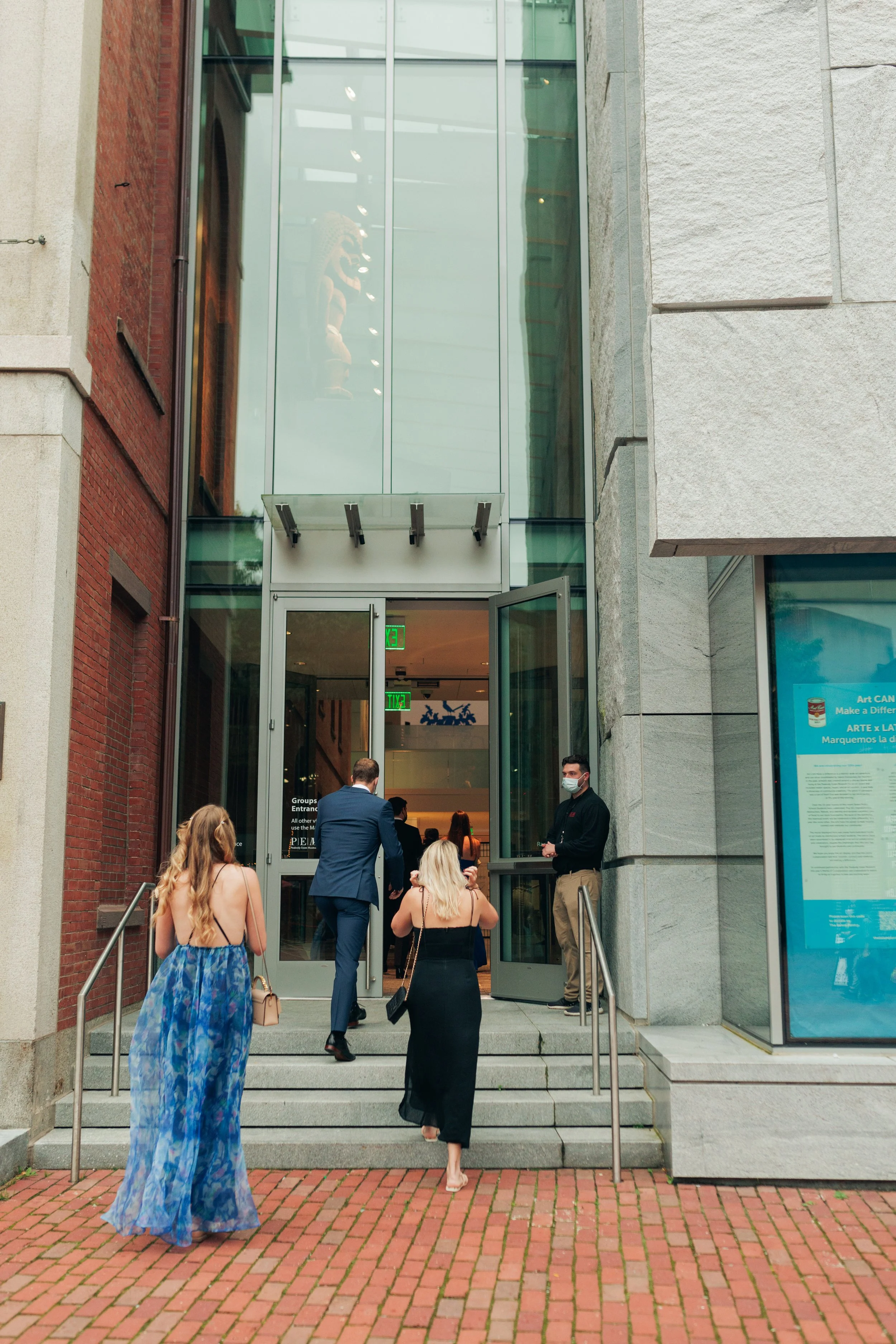 People entering a building through glass doors on a city sidewalk, some dressed in formal attire, some wearing masks.