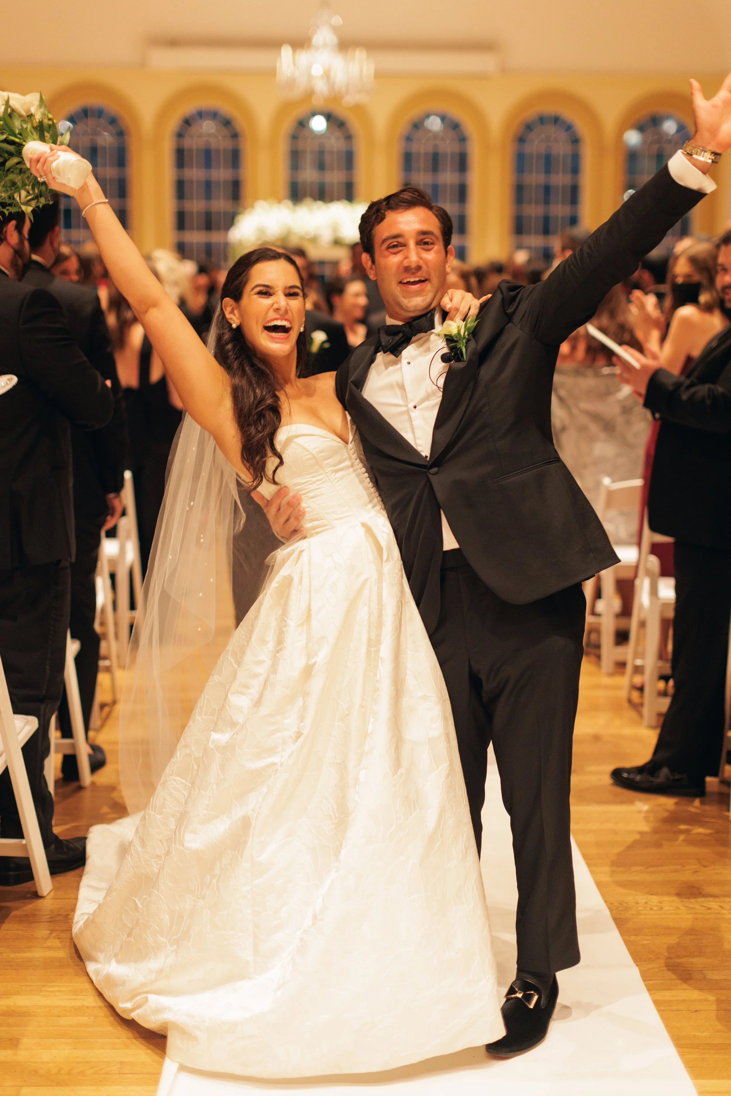 Happy bride and groom dancing at their wedding reception in a decorated hall with large windows and chandelier.