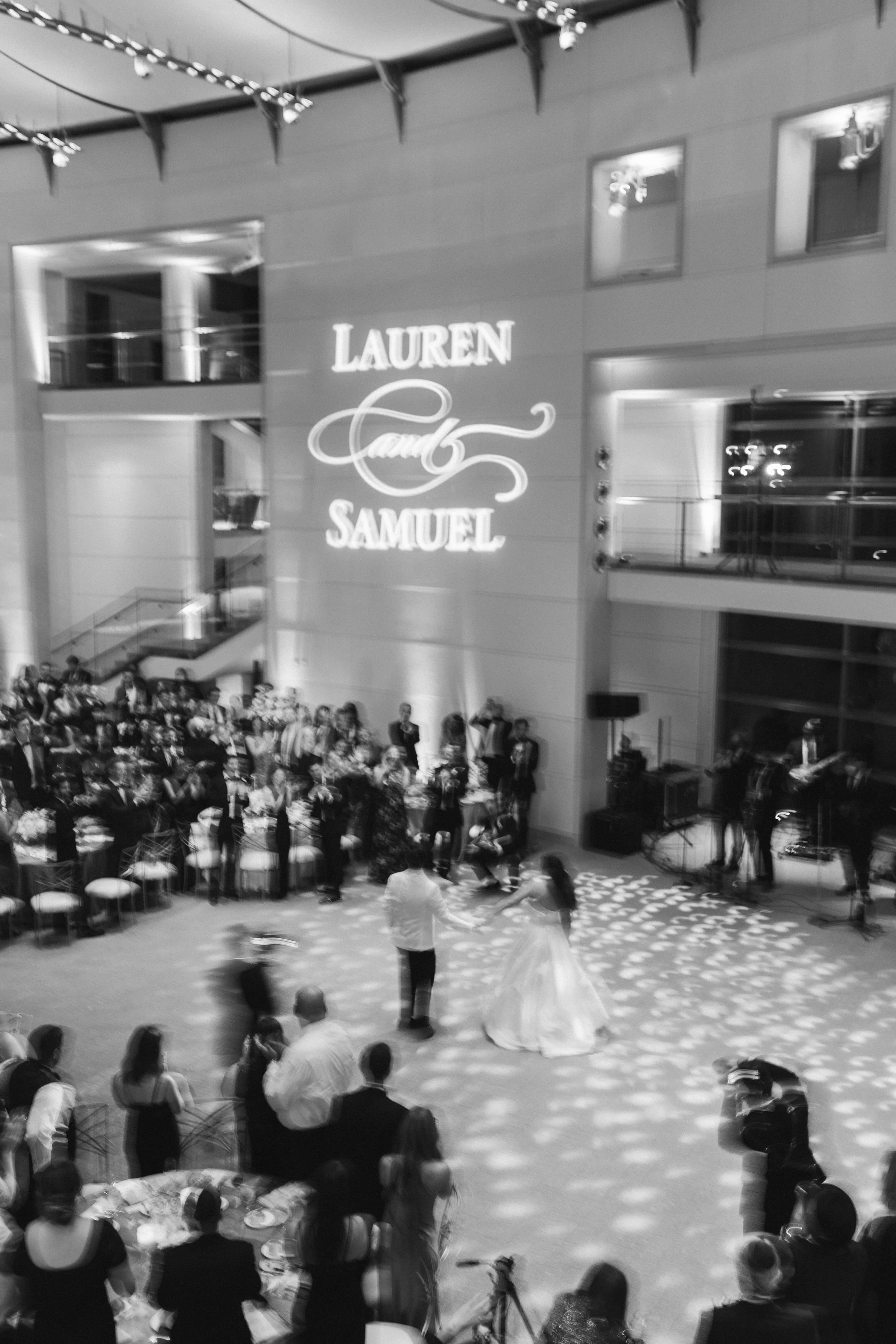 A wedding dance event in an indoor venue with a large crowd. There is a couple dancing in the foreground, with the bride in a long white gown and the groom dressed formally. The background shows a stage area with a projected sign reading 'Lauren and 