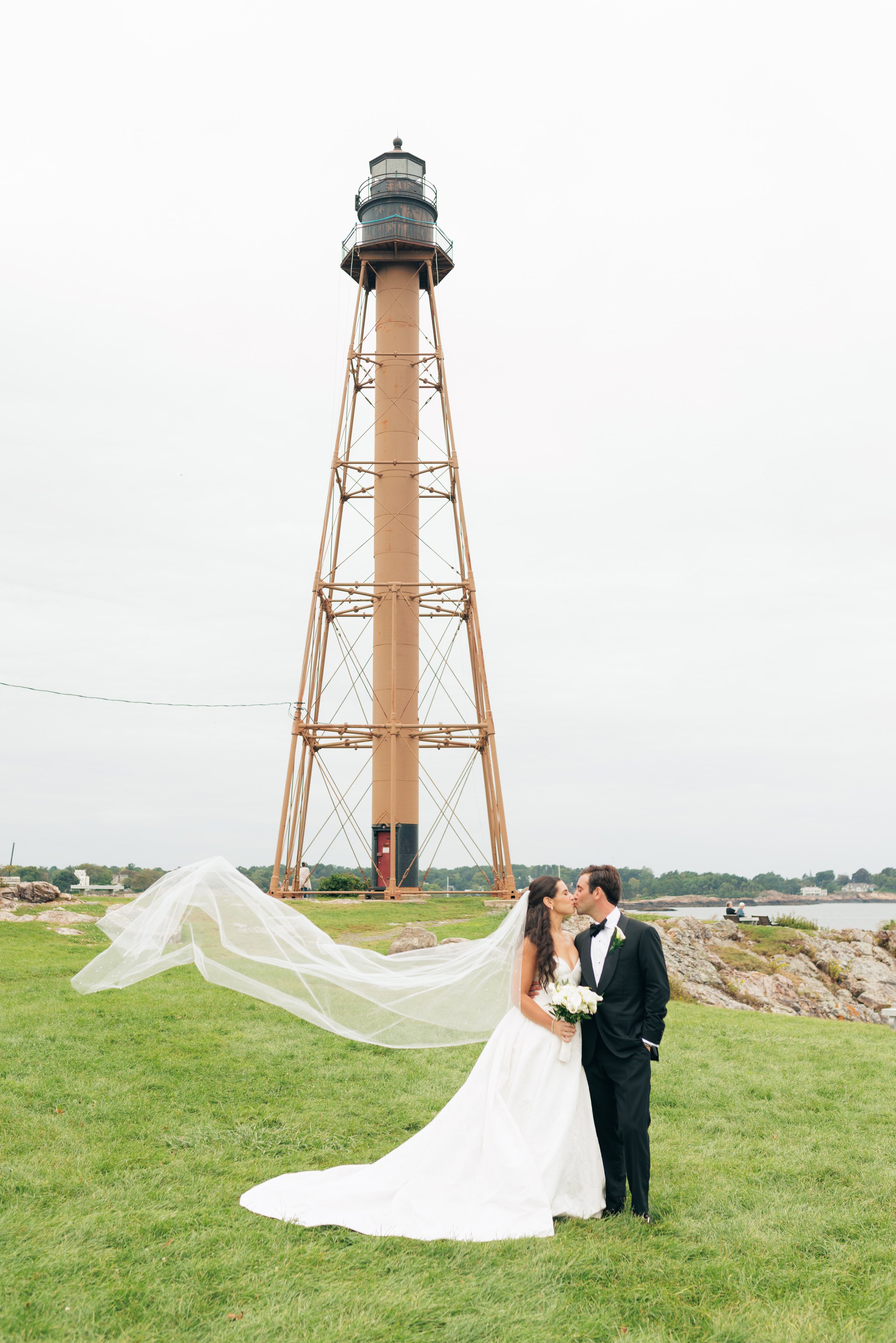 A newlywed couple kissing outdoors near a lighthouse, with the bride holding a bouquet and a white veil flowing on the grass.