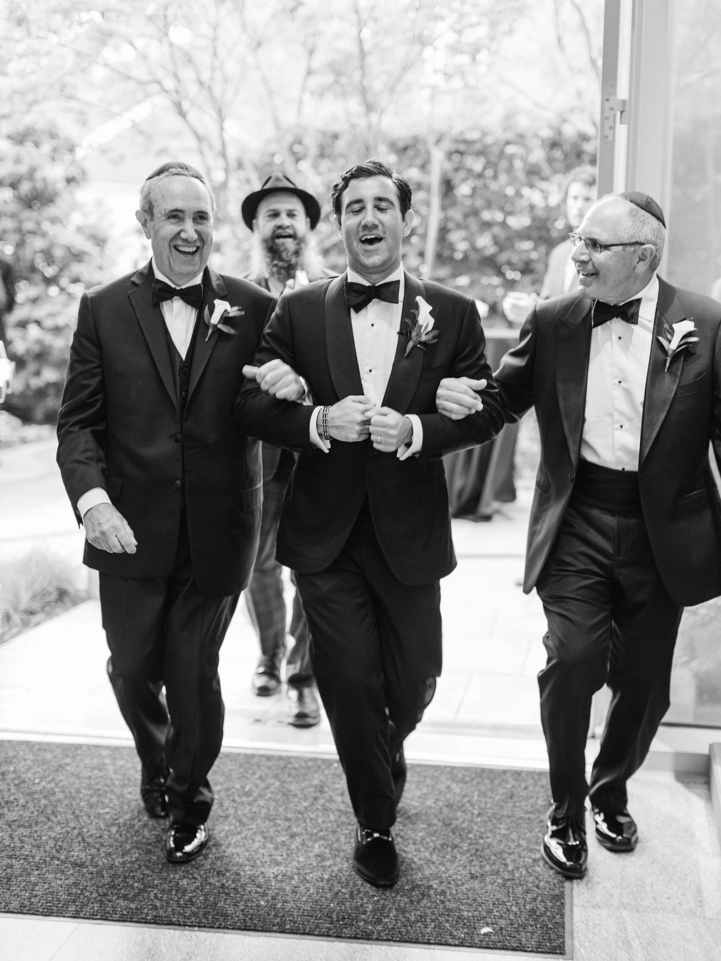 Black and white photo of four men in tuxedos and bow ties, walking together outdoors. The man in front appears to be singing or speaking, with men on either side and behind him sharing in the moment, smiling and engaging with him.