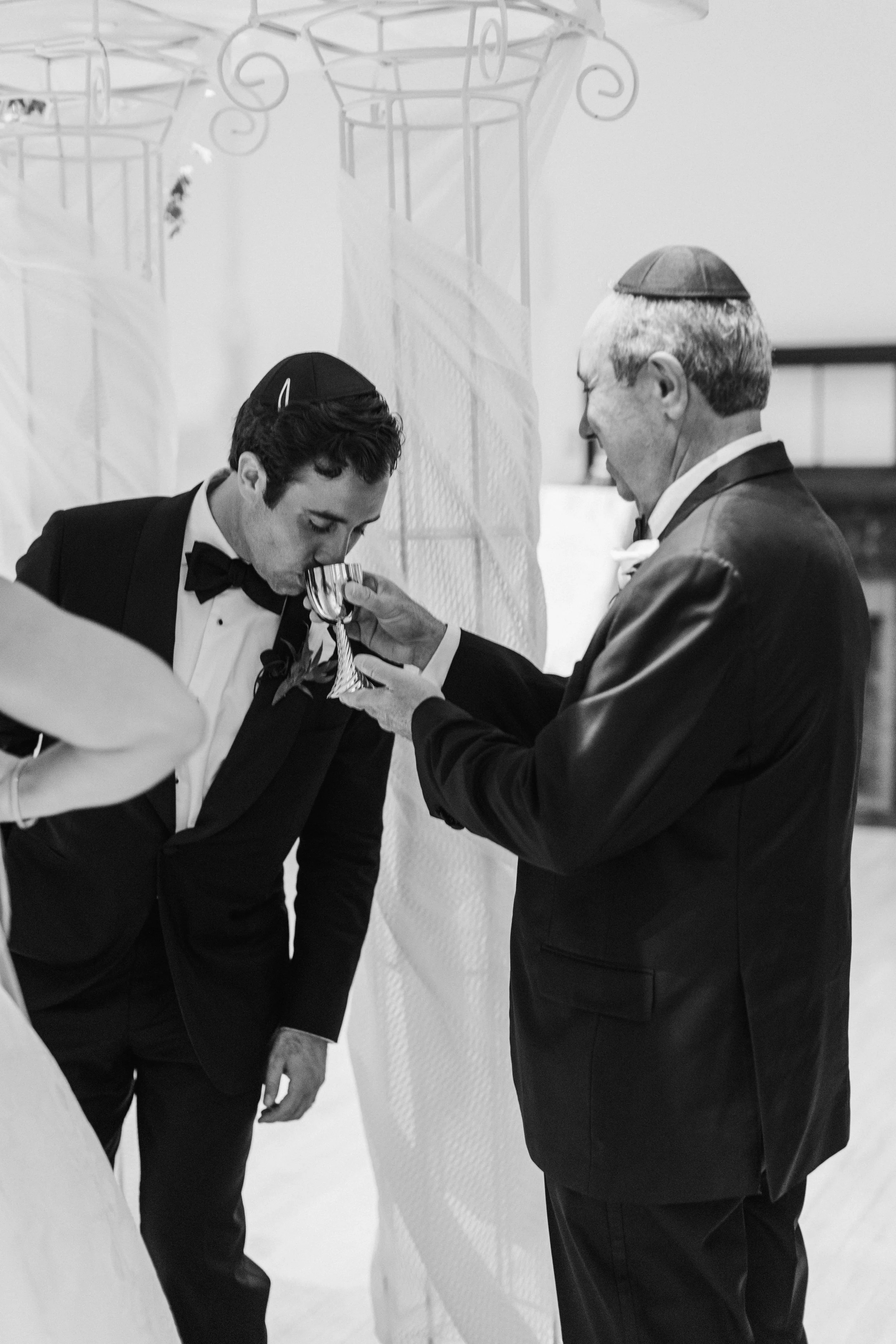 A young man dressed in a tuxedo with a bow tie is leaning forward, with a hand on his knee and head bowed, while an older man dressed in a suit and bow tie is helping him to drink from a goblet during a social event.