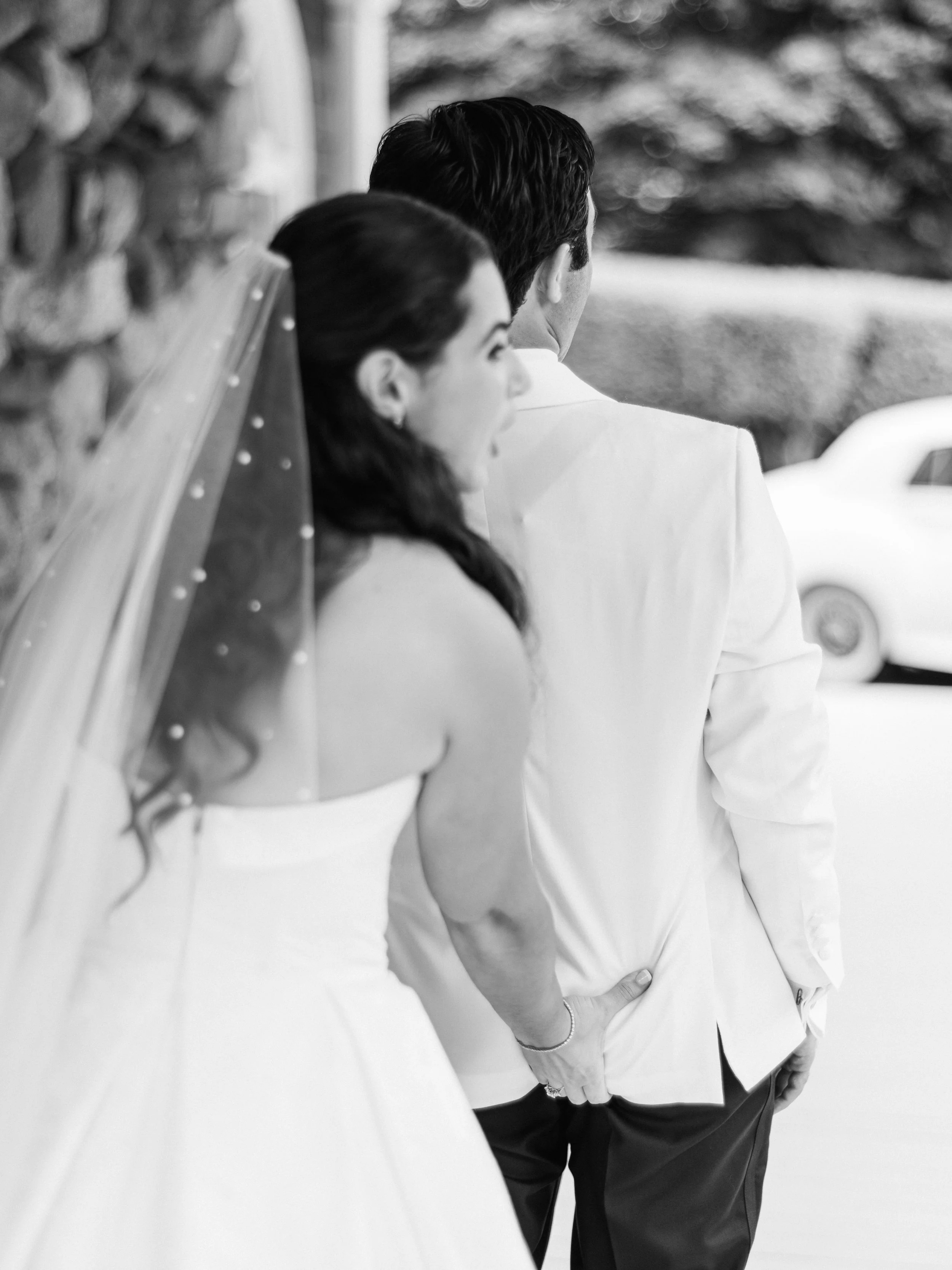 Black and white photo of a couple standing close together, facing away from the camera, with the woman smiling and leaning towards the man, who is wearing a white blazer and dark pants, outdoors with blurred trees and a vehicle in the background.