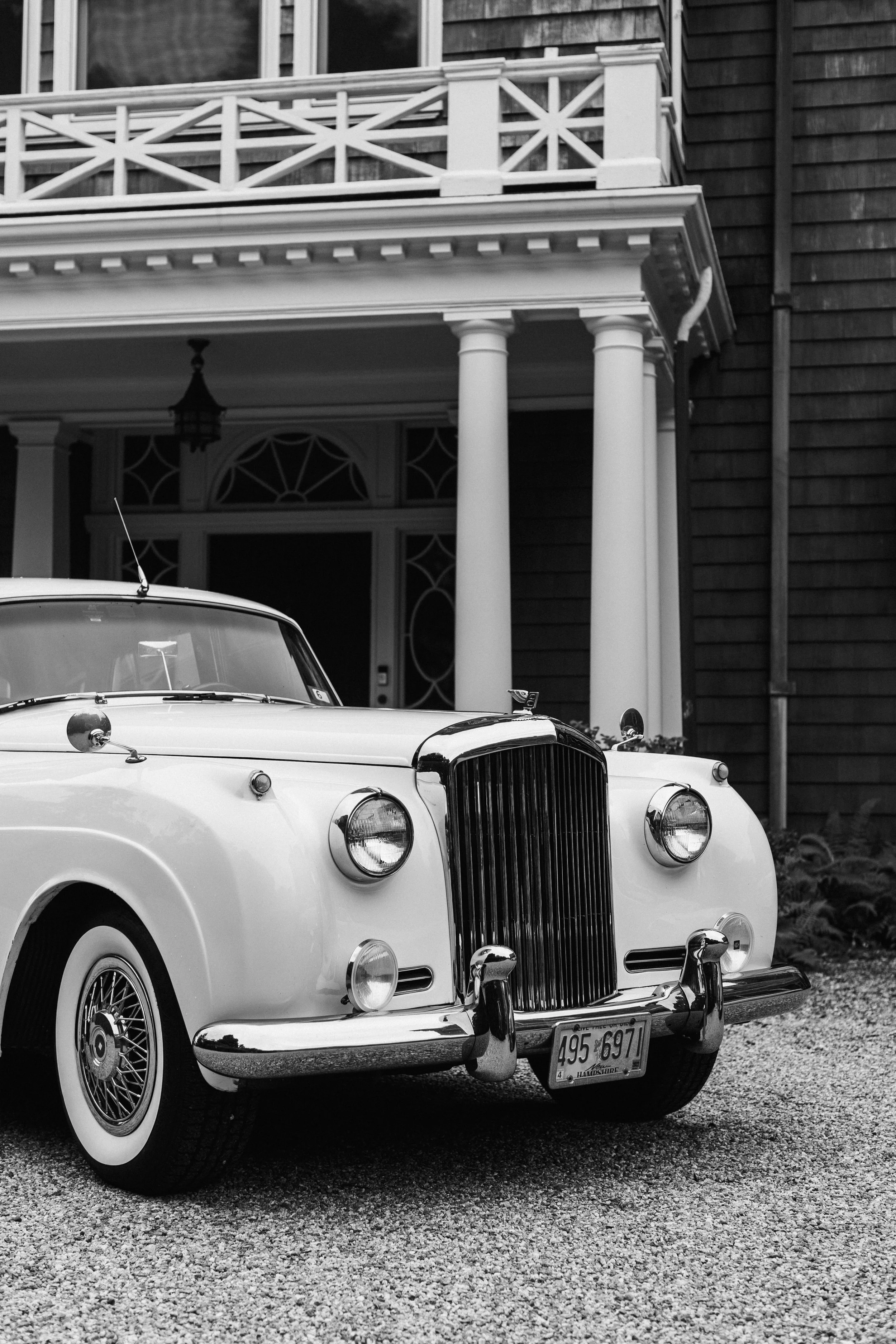 Black and white photo of a vintage white car parked in front of a house with a porch and columns, and wooden siding. The car has a classic design with a grille, headlights, and a Maryland license plate.