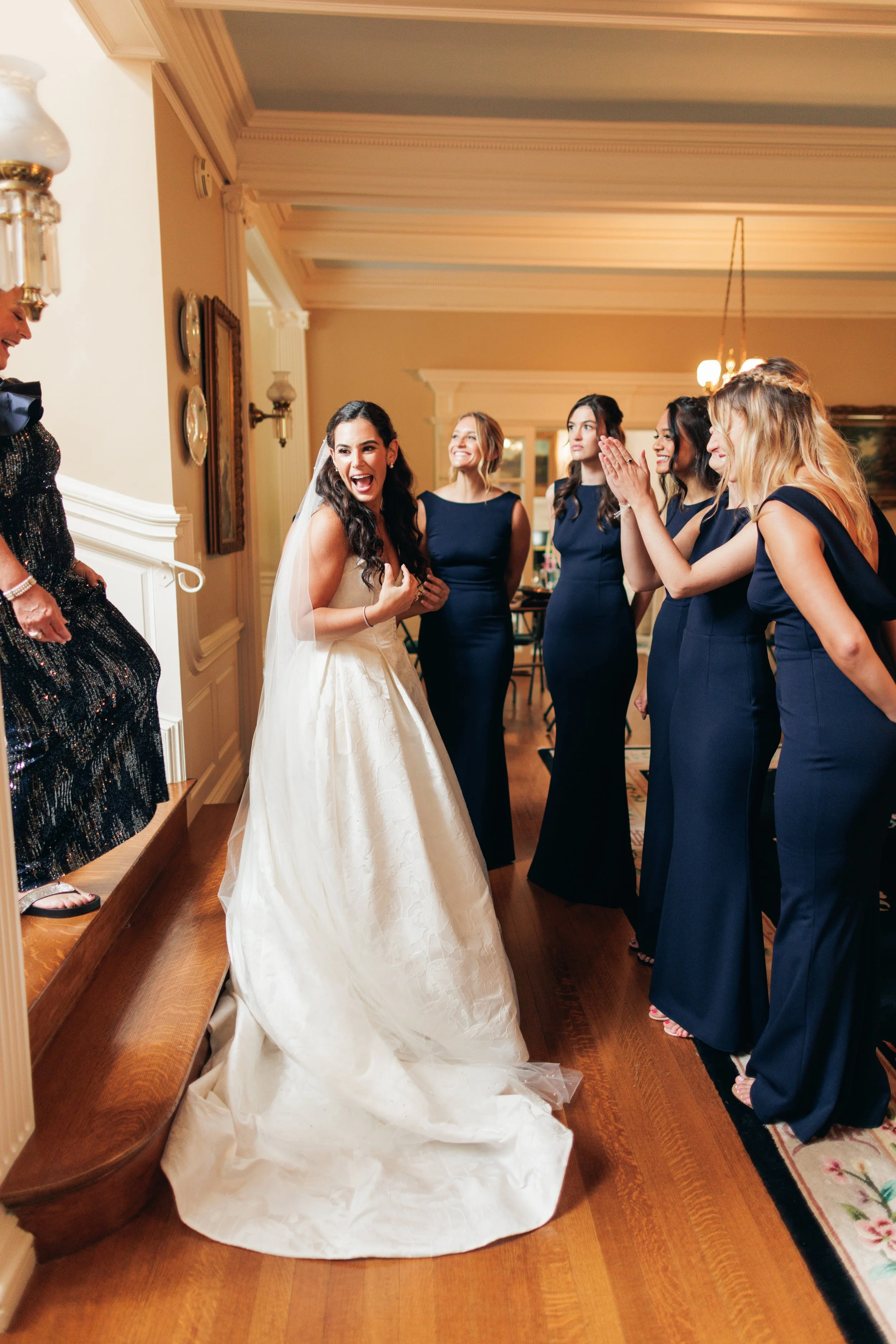 A bride in a white wedding gown with a train, smiling and laughing, surrounded by five women in navy blue dresses, in an elegant indoor setting with warm lighting and wooden flooring.