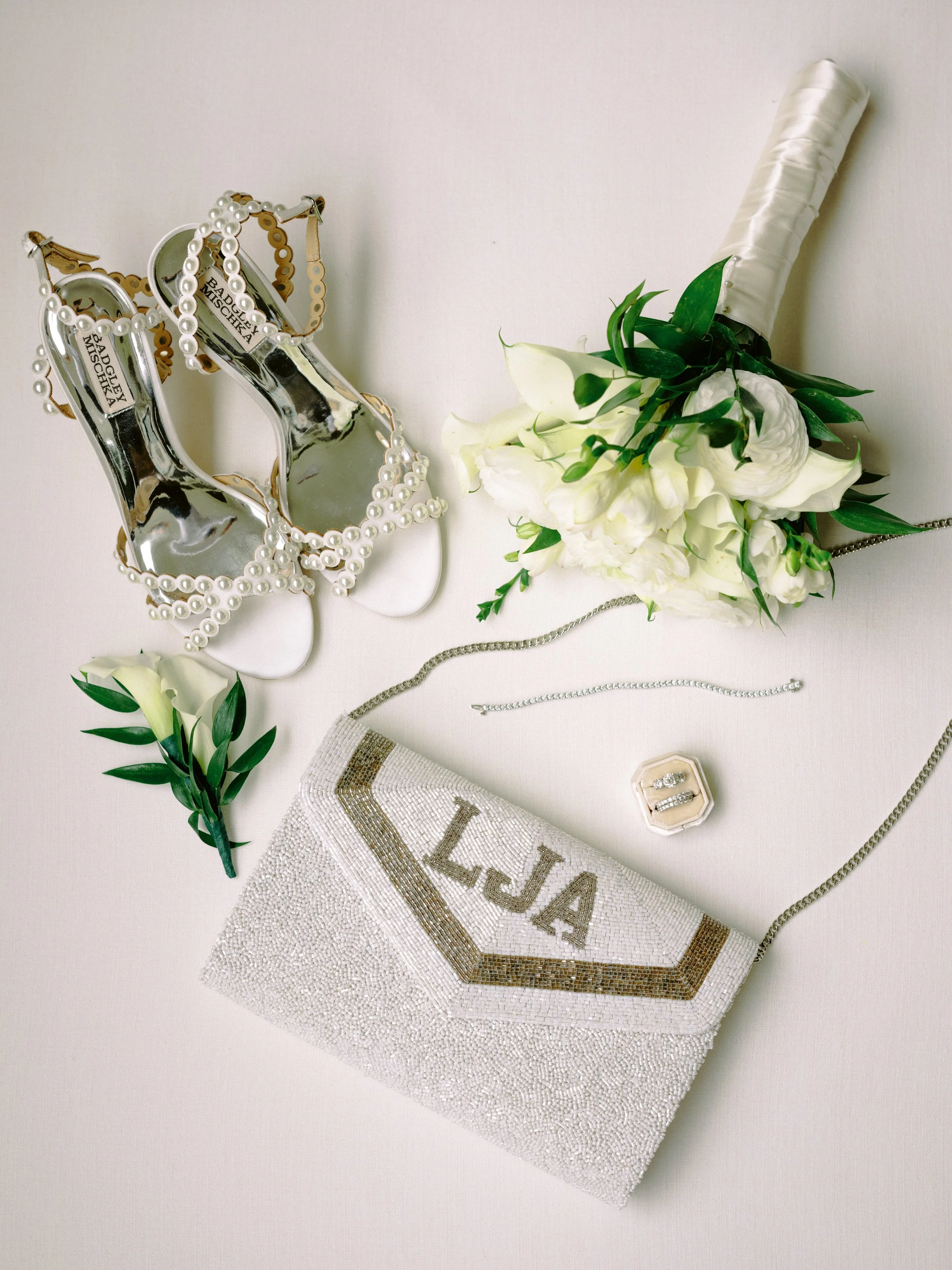 A pair of high heel shoes with pearl embellishments, a white floral boutonniere with green leaves, a matching white purse with gold embroidery, a silver necklace, a jewelry box with rings, and a white curtain with a gold border.
