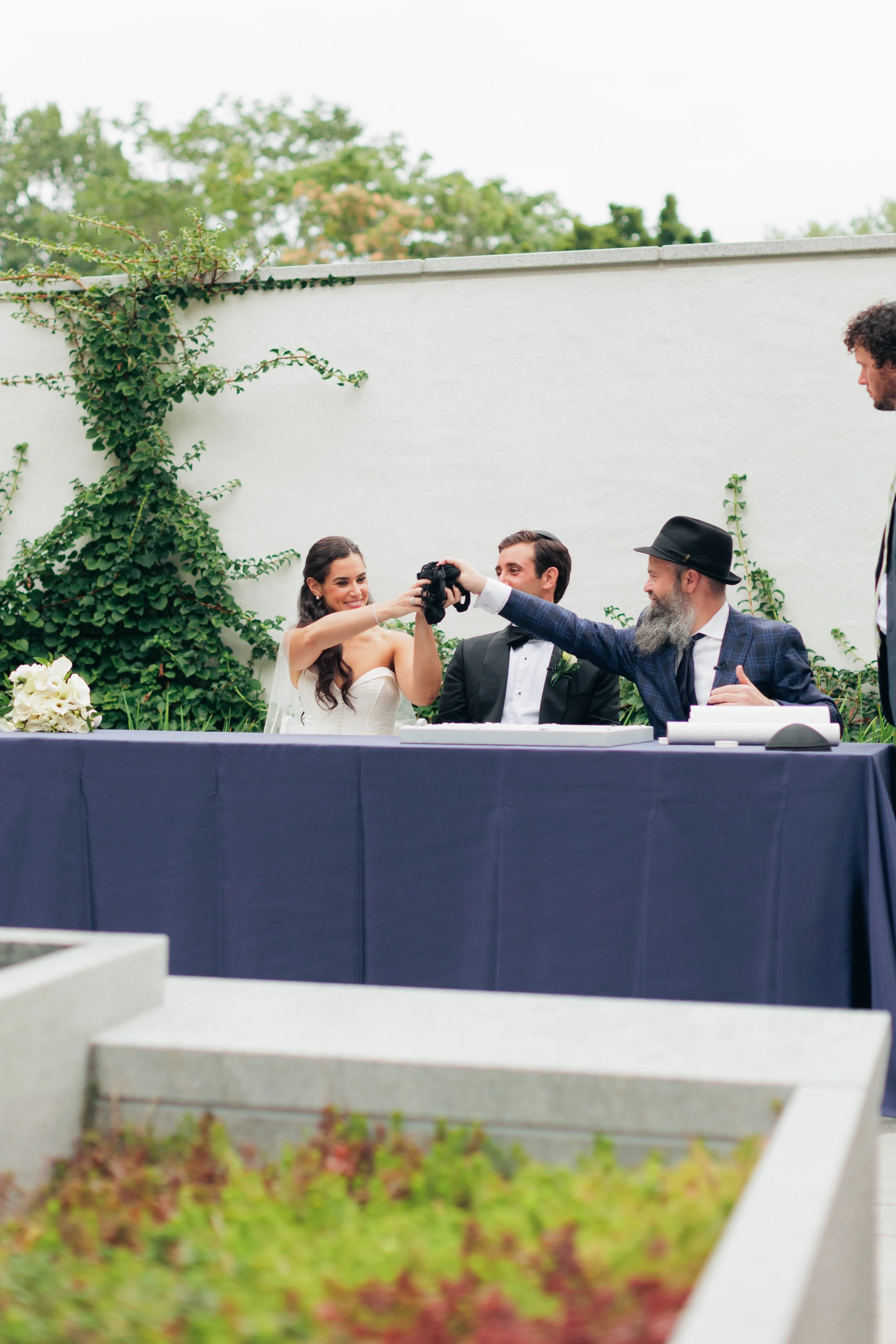 A bride and groom sitting at a table with three men at an outdoor wedding reception, with a white wall and trees in the background.