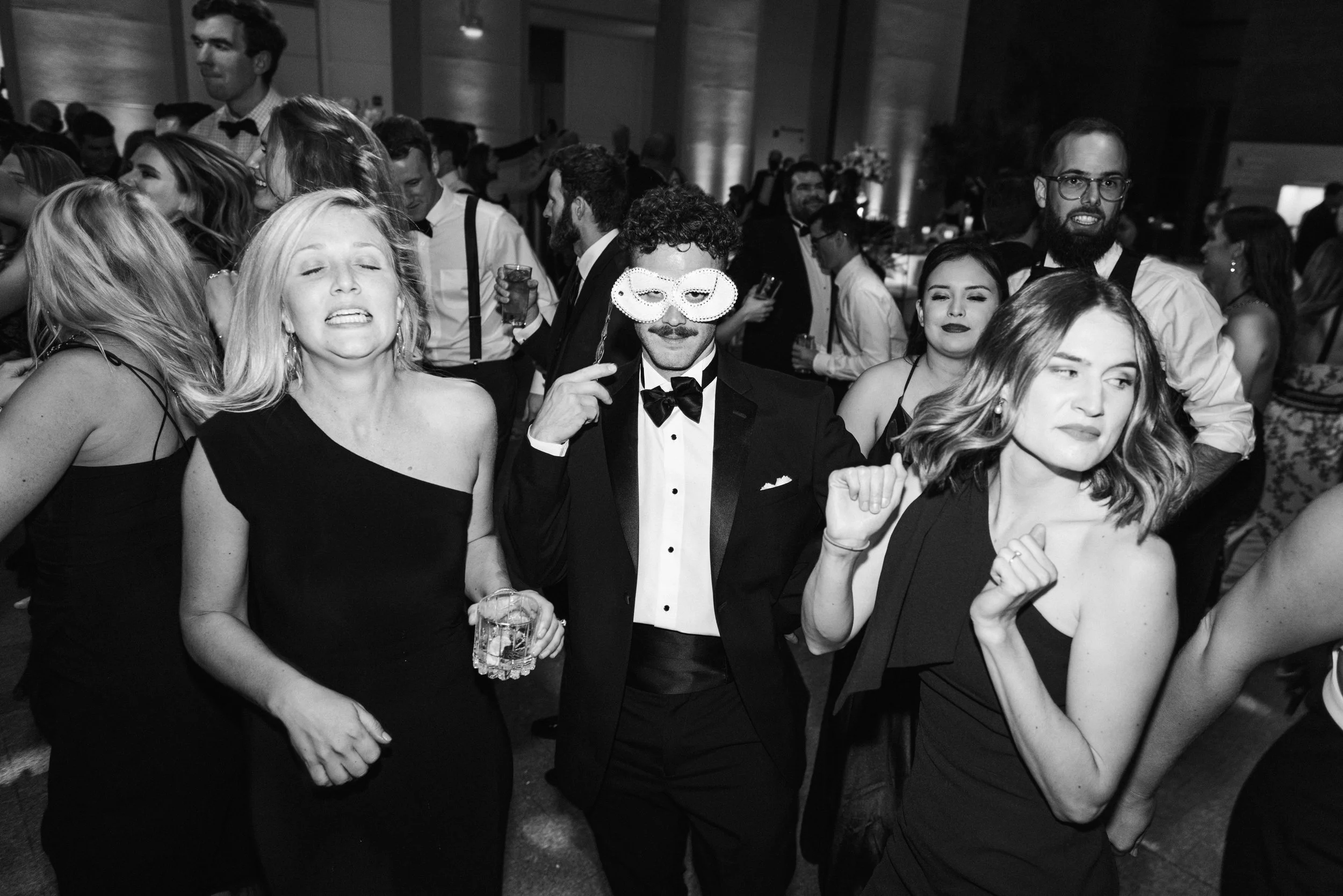 People at a formal event or party, dressed in elegant attire, with a man in a tuxedo wearing a novelty mask, holding a drink, surrounded by smiling women and other guests.