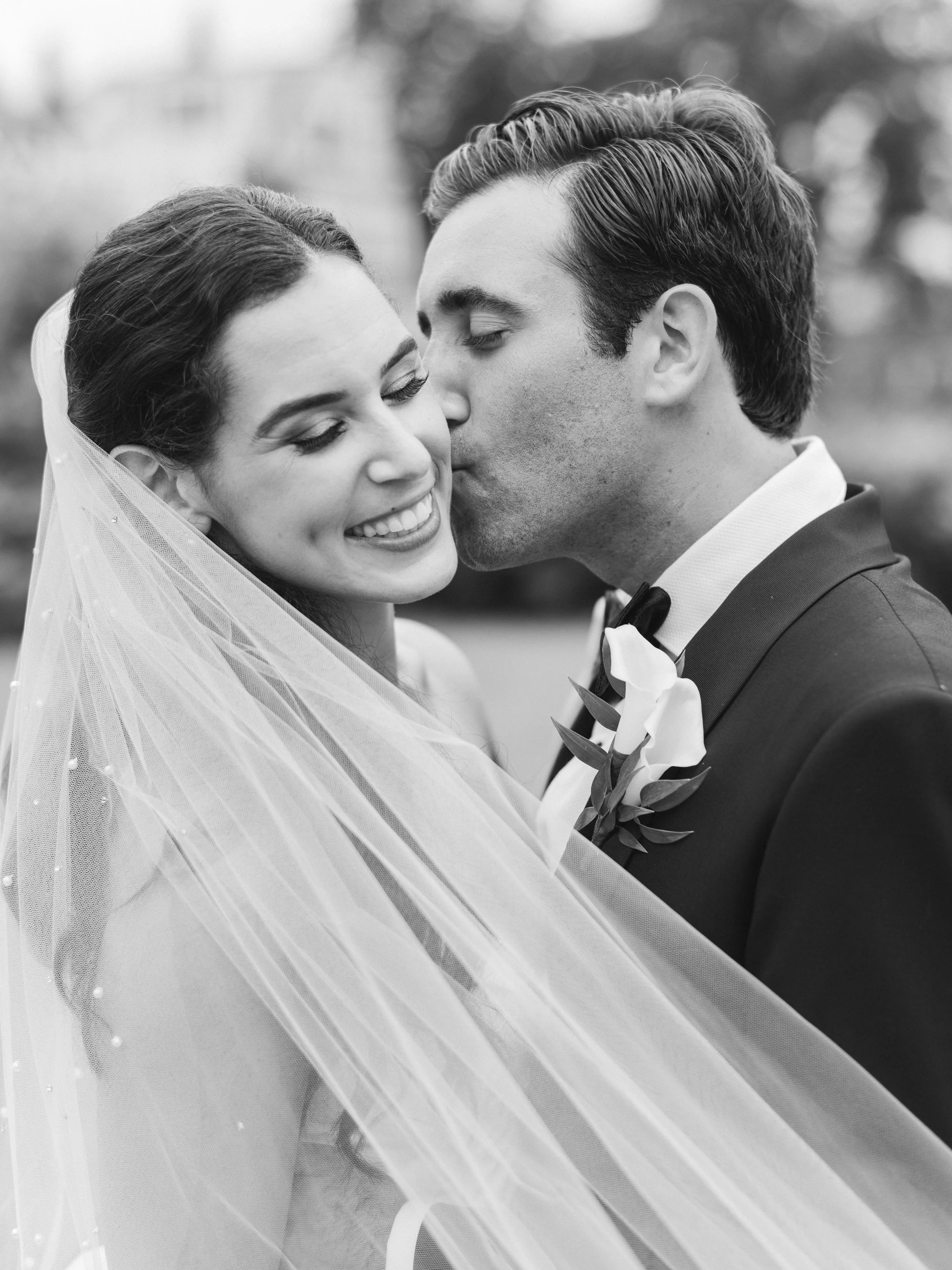 Black and white photo of a bride and groom close together, smiling and about to kiss, with the bride wearing a veil and the groom in a tuxedo.