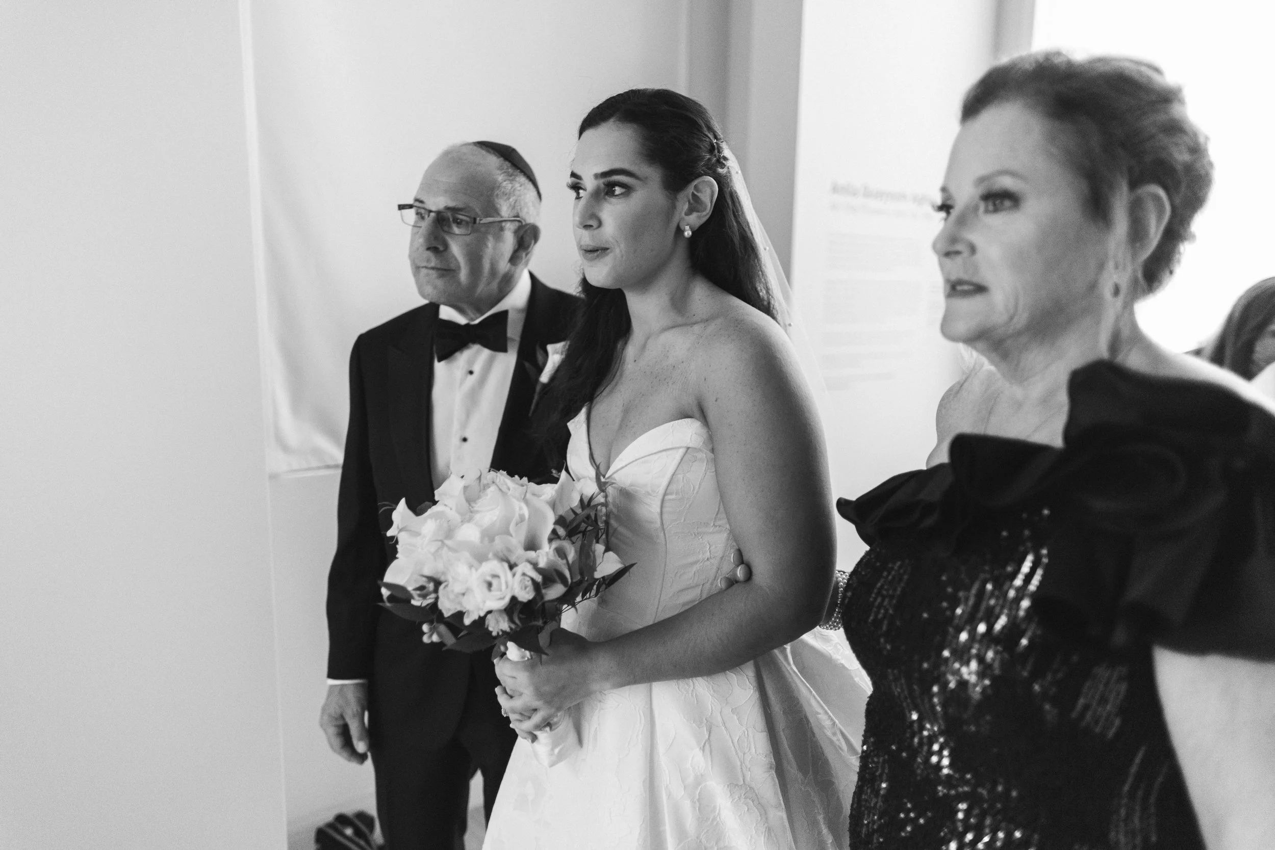 A bride holding a bouquet of white roses standing next to two women, one man, all dressed formally at a wedding ceremony.