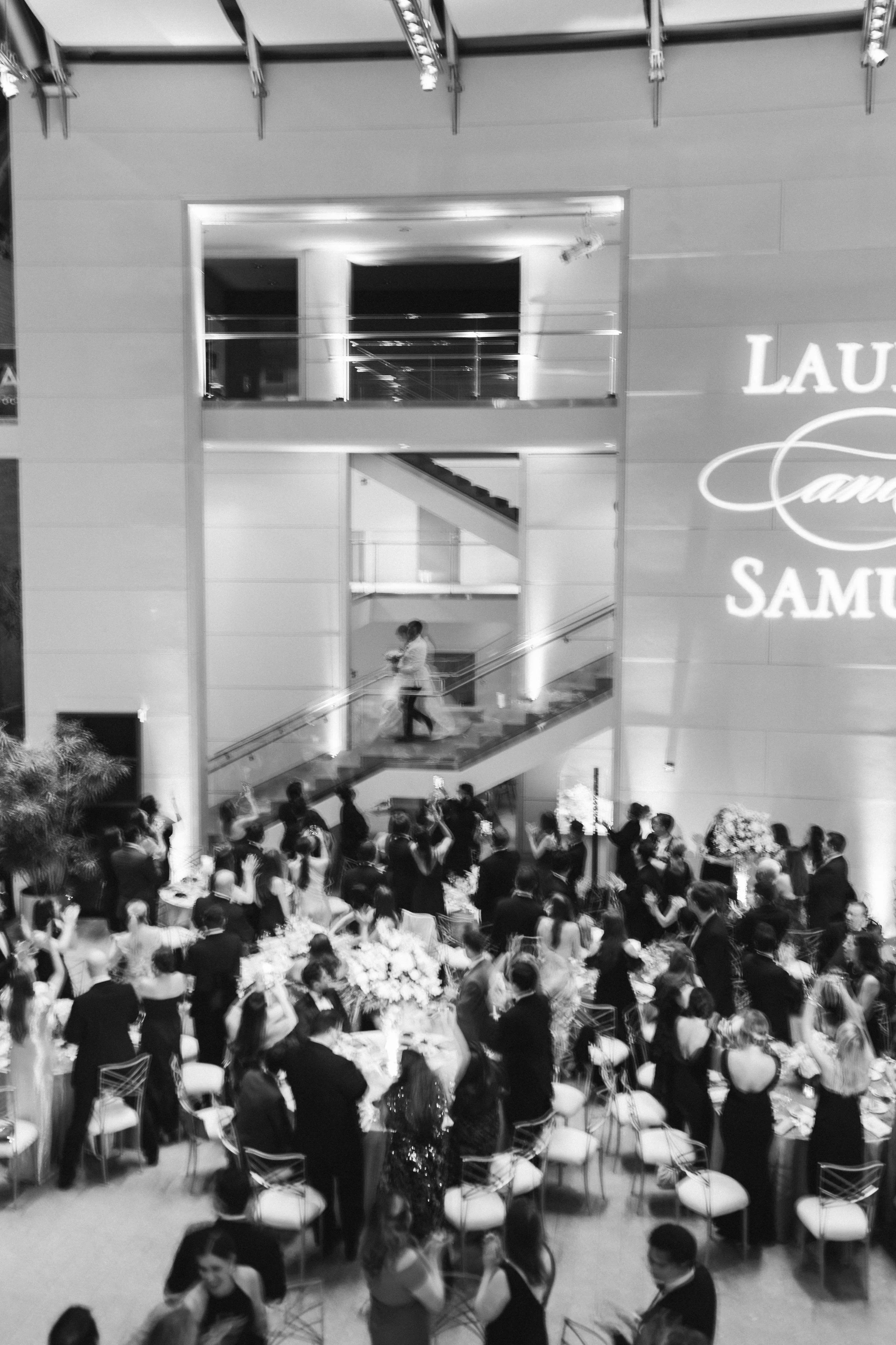 A large gathering of people in an elegant indoor space, with round tables decorated with floral arrangements, and guests dressed in formal attire. Multiple levels are visible, including a balcony and staircase, and there is a large illuminated sign o