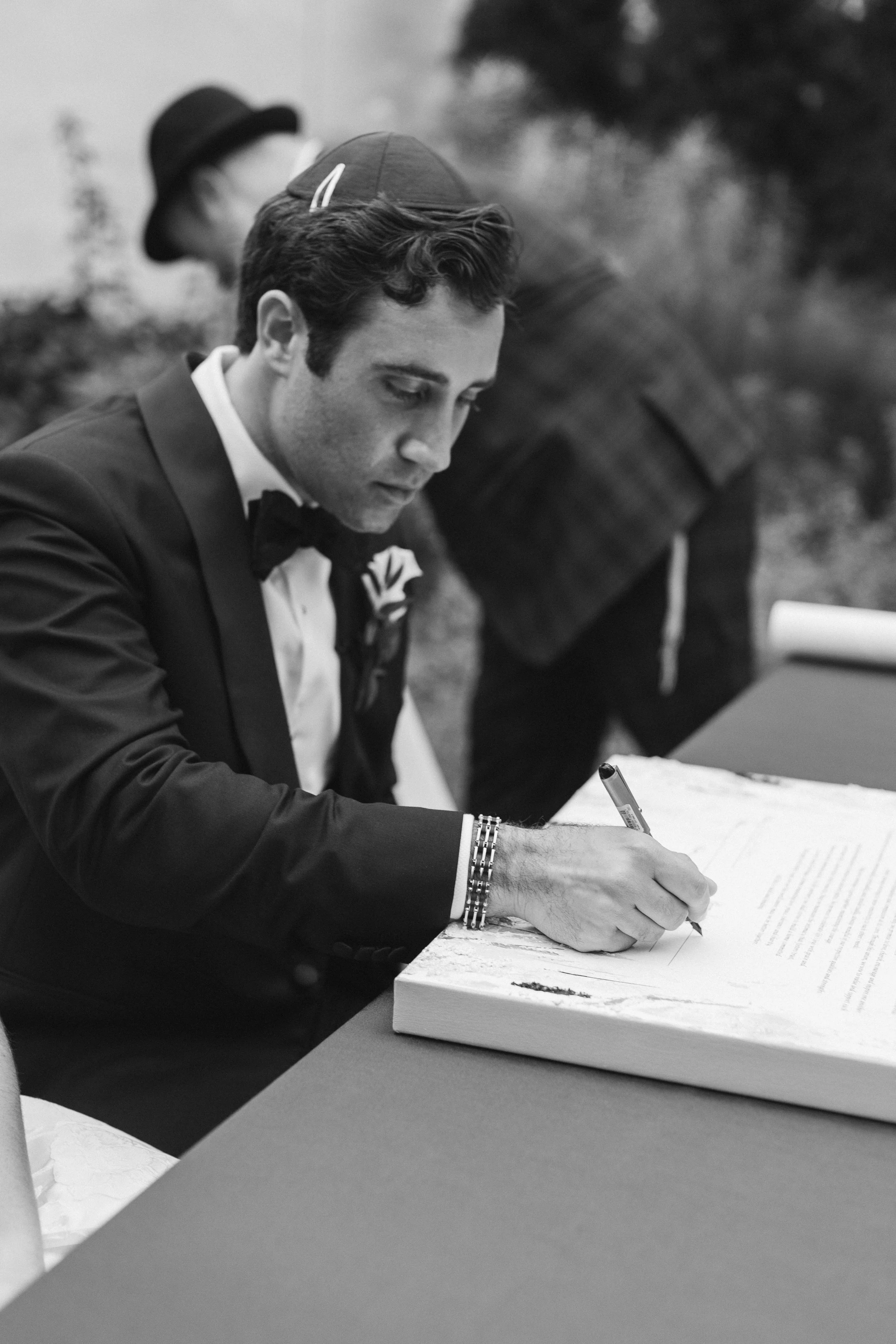 A man in a tuxedo signing a document, framed in black and white, with another person in the background. The man is wearing a bow tie and has a stack of bracelets on his wrist.