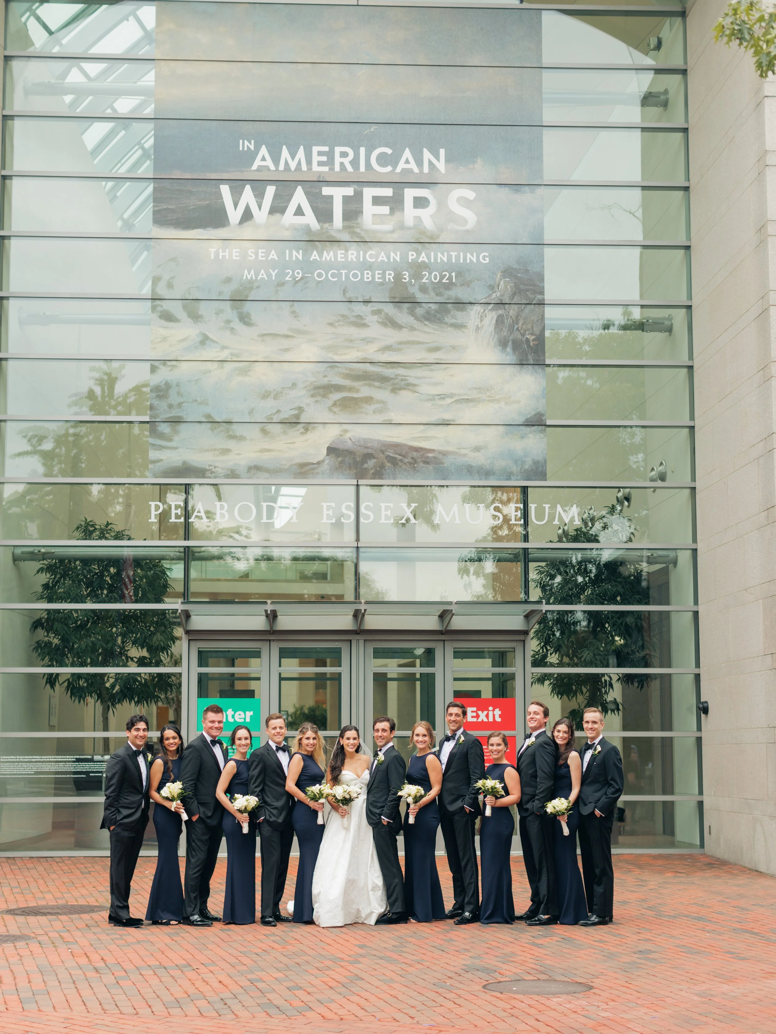 A group of twenty people, including a bride and groom, standing in front of the Peabody Essex Museum entrance during a wedding celebration. They are dressed semi-formally with women in matching navy dresses and men in suits. The background shows the 