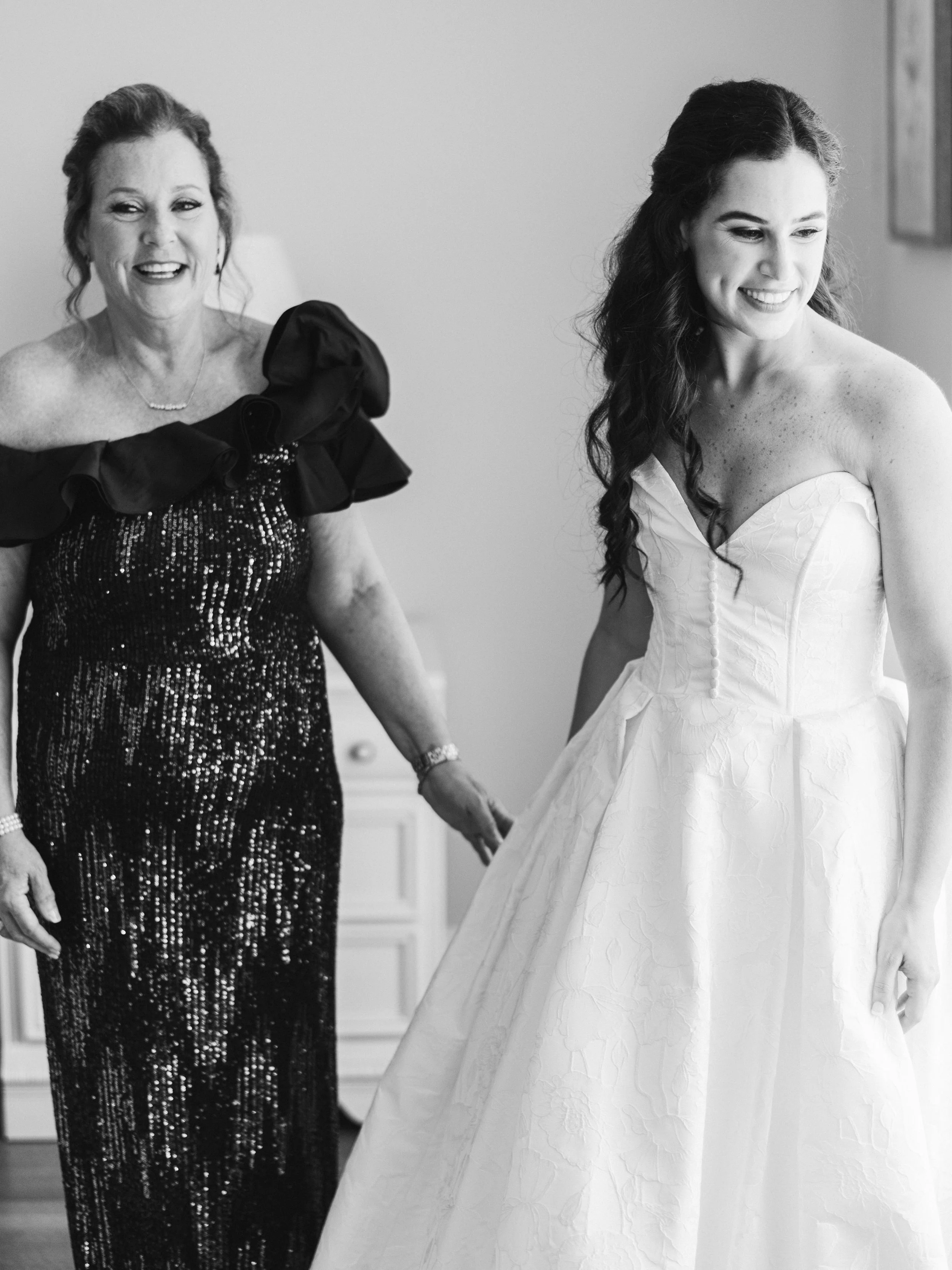A woman in a wedding dress with long, dark wavy hair smiling, being assisted by another woman in a formal dress.