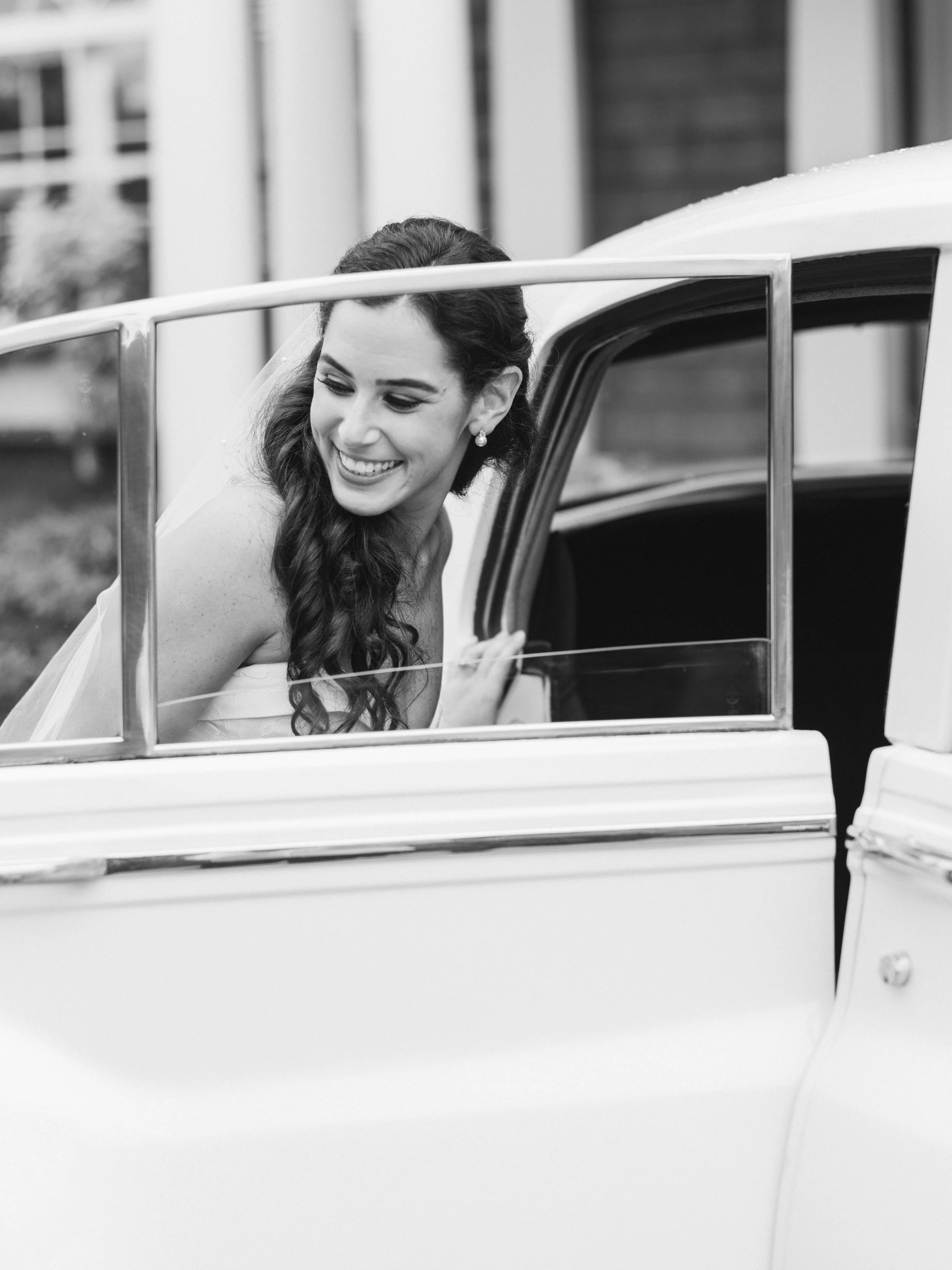 A smiling woman with long, curly hair and pearl earrings leaning out of the window of a vintage white car.