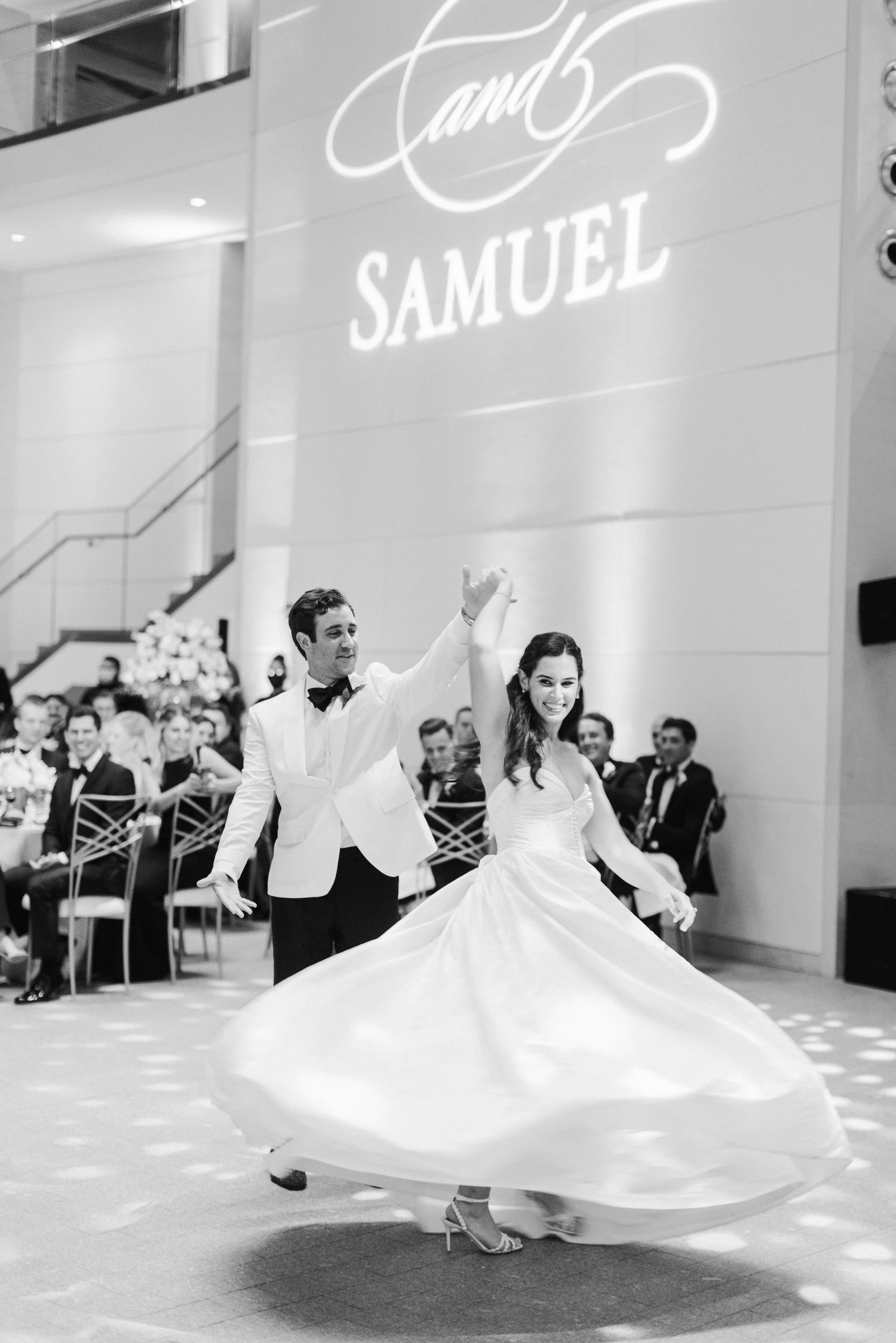 A couple dancing at a formal event, with the woman twirling in a long, flowing gown and heels, and the man in a tuxedo. They are in an indoor venue with a large audience and a sign that says 'Grand Samuel' in the background.
