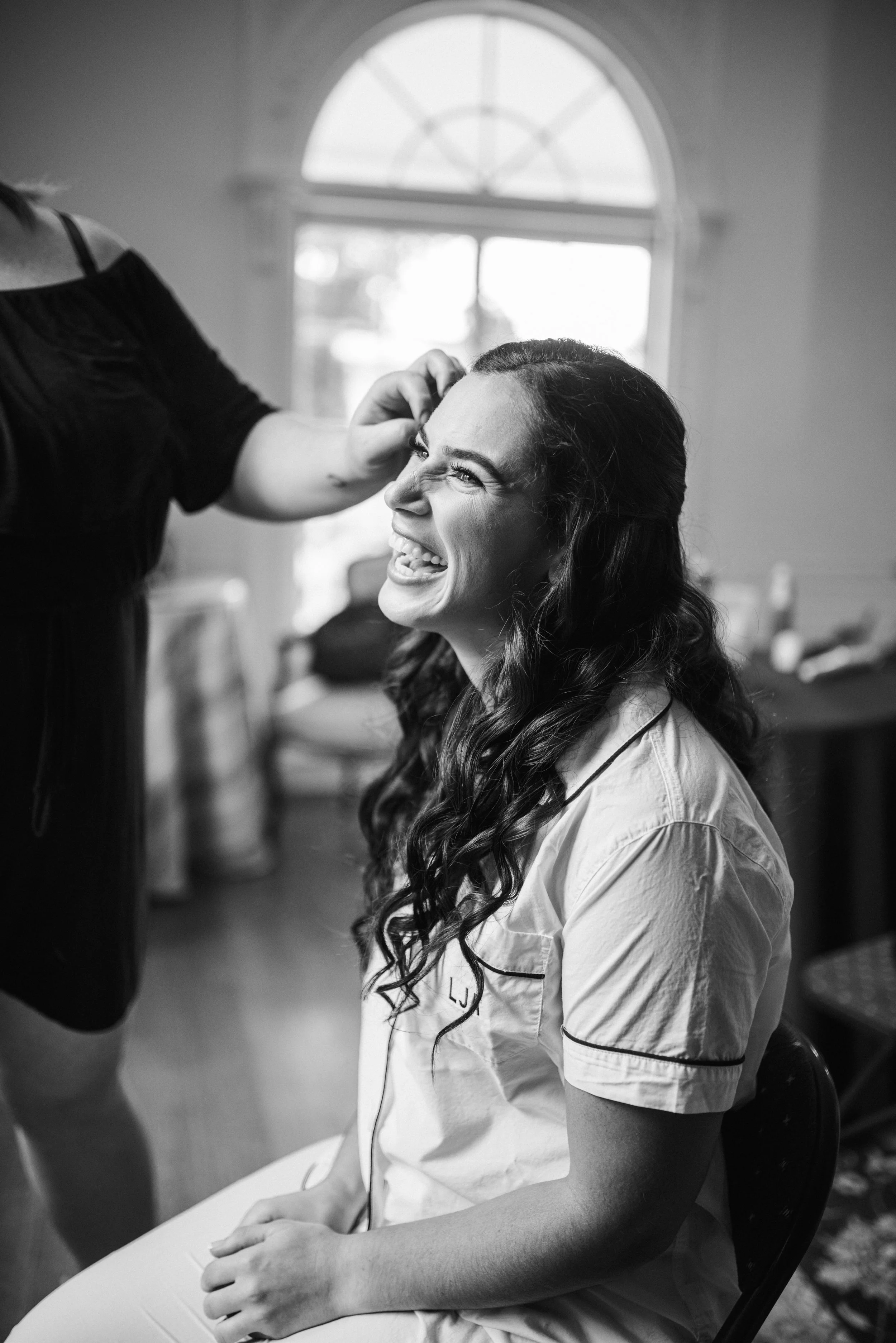 Black and white photo of a woman sitting in a chair, smiling and laughing while getting her hair styled by a stylist.