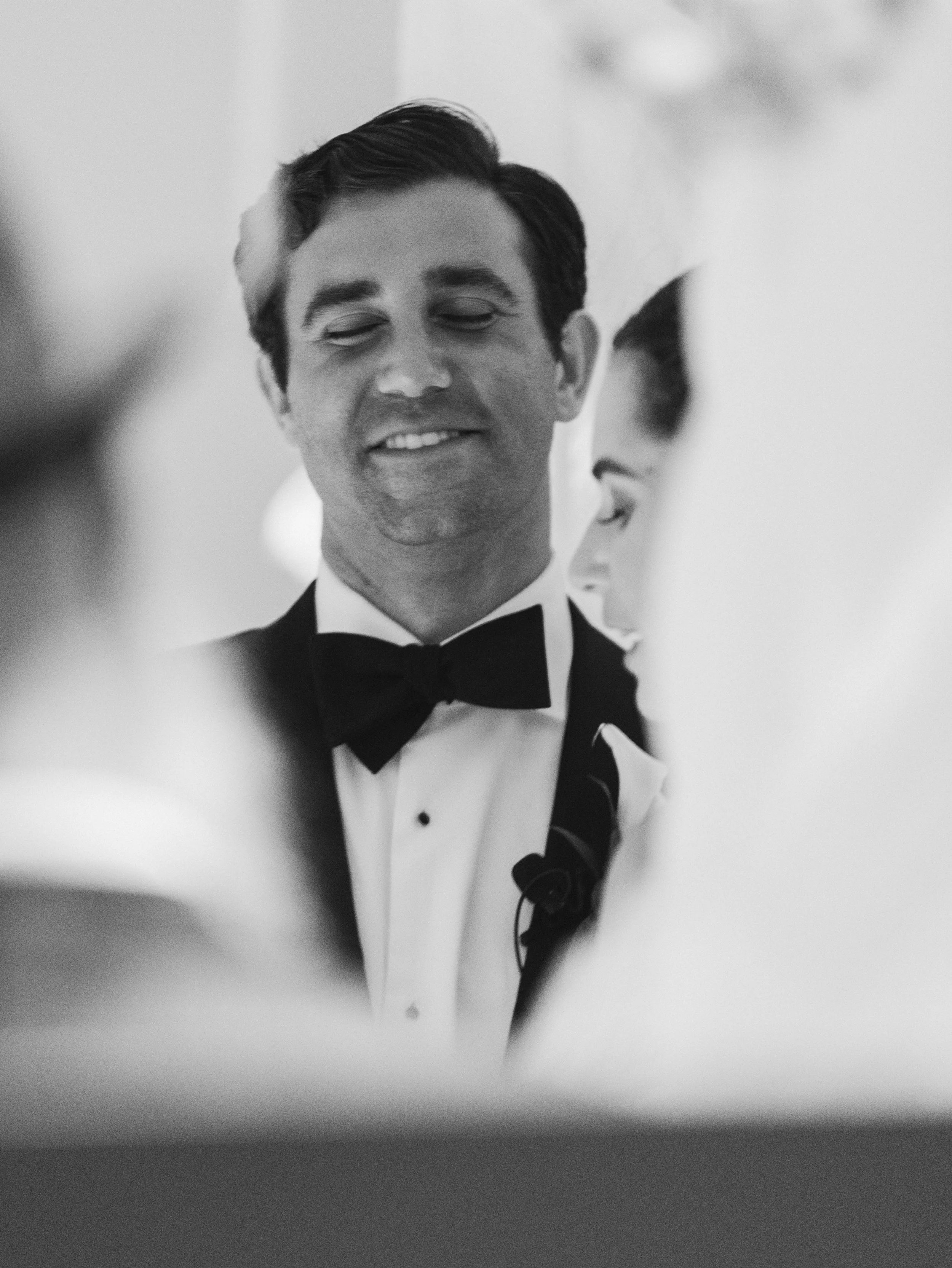Black and white photo of a man in a tuxedo with a bow tie, smiling with closed eyes, near another person's face. The focus is on his face and tuxedo.