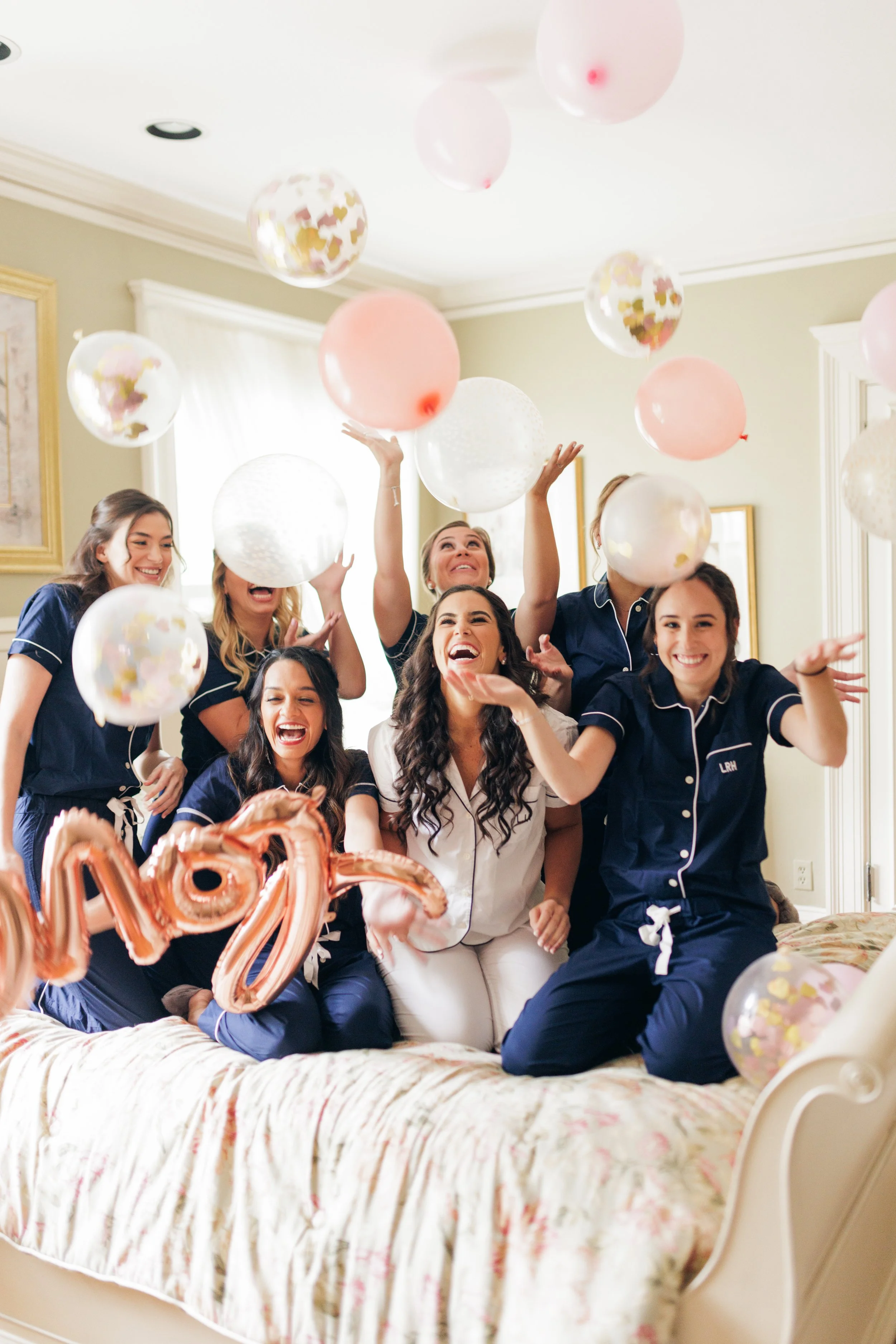 Celebrating New Year's Eve in a bedroom with several young women wearing navy pajamas, laughing and playing with balloons and rose gold '2023' balloons, with pink and white balloons floating, party decorations, and a bed with a floral bedspread.