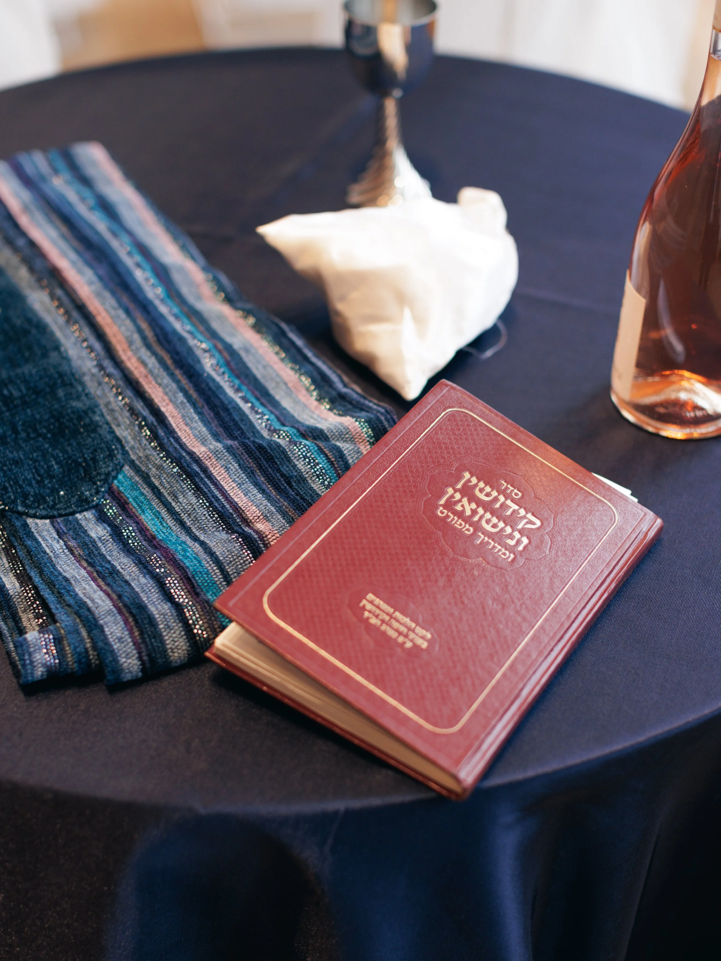 A black round table with a colorful striped cloth, a white conch shell, a red book with Hebrew writing, a small pinkish-brown glass bottle, and a dark cup in the background.