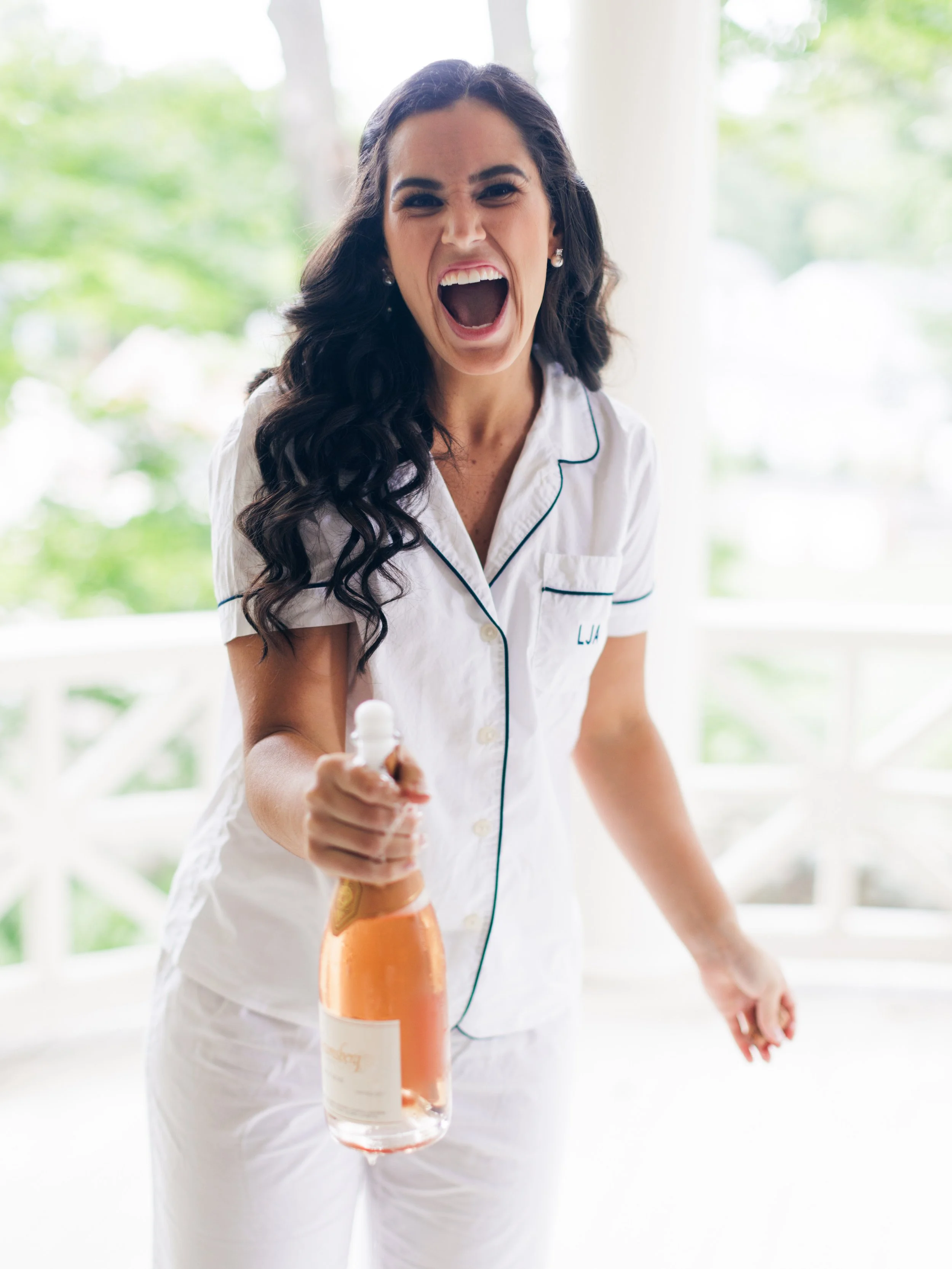 A woman in white pajamas with black piping, laughing and holding a bottle of orange juice, standing in a bright room with a large window and greenery outside.
