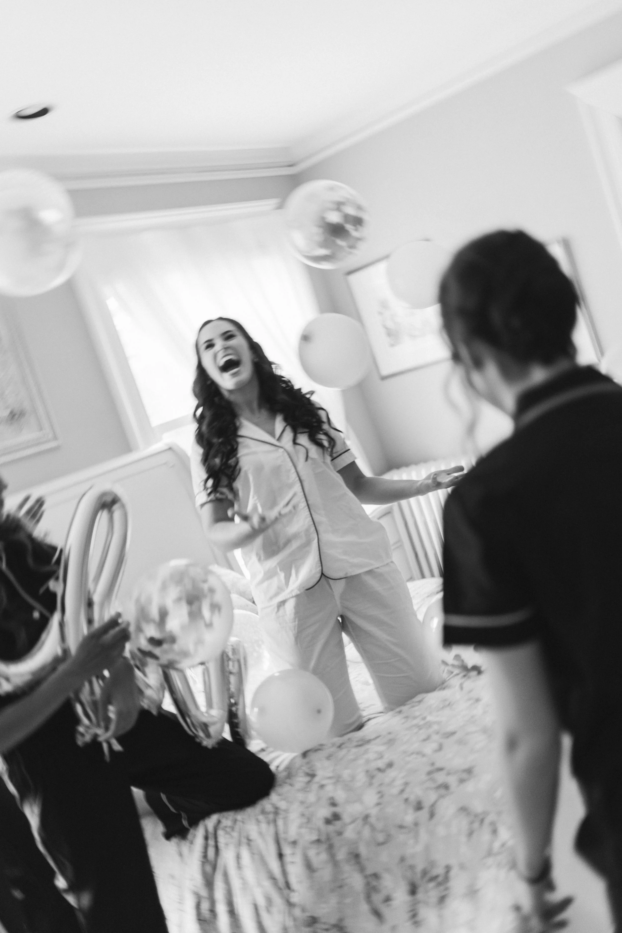 A woman dressed as a nurse laughing, with balloons and a person holding a decorated cake in a hospital room.