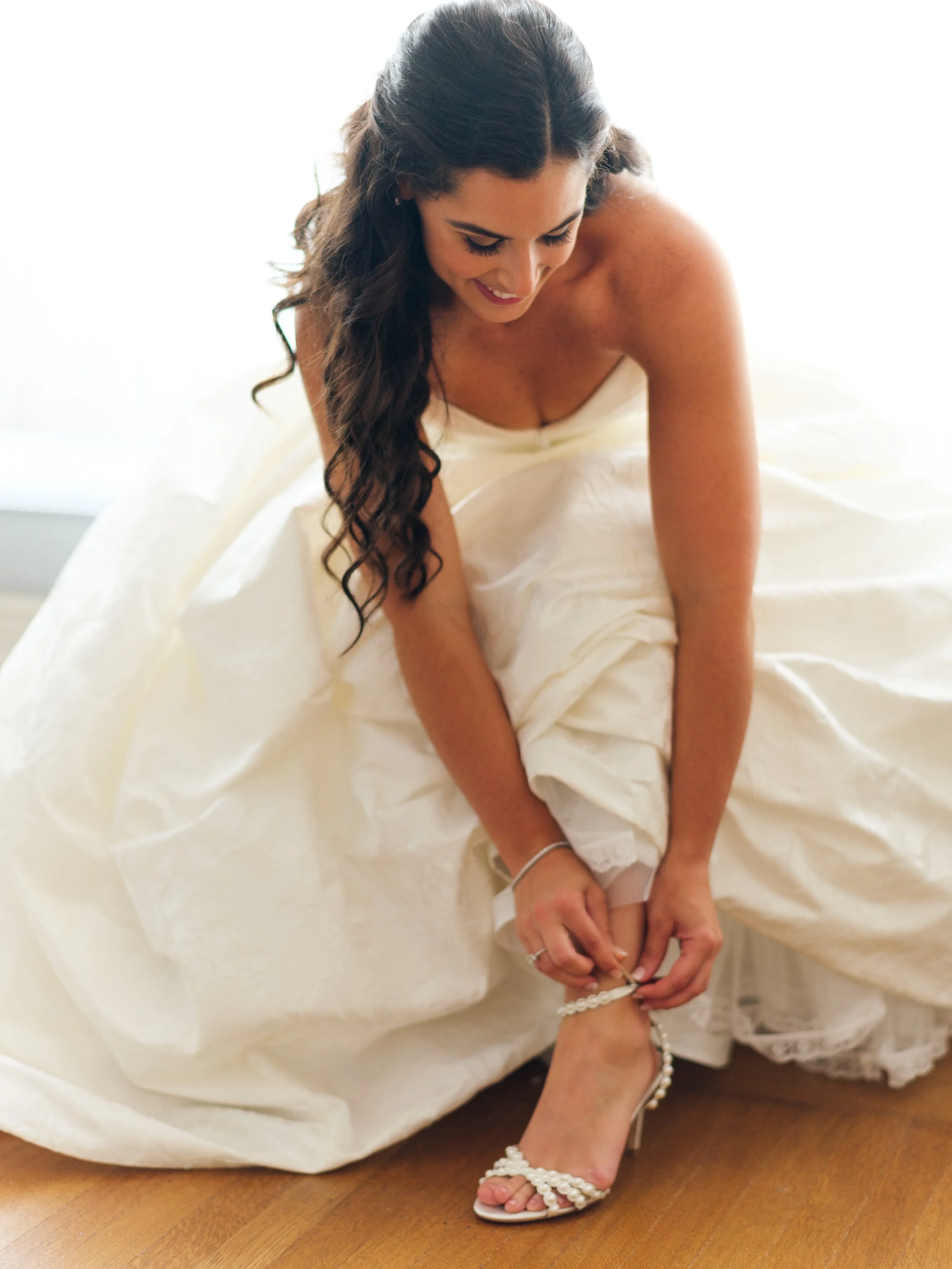 A woman, dressed in a wedding gown, sitting on a bed and helping someone put on a pearl-strap sandal.