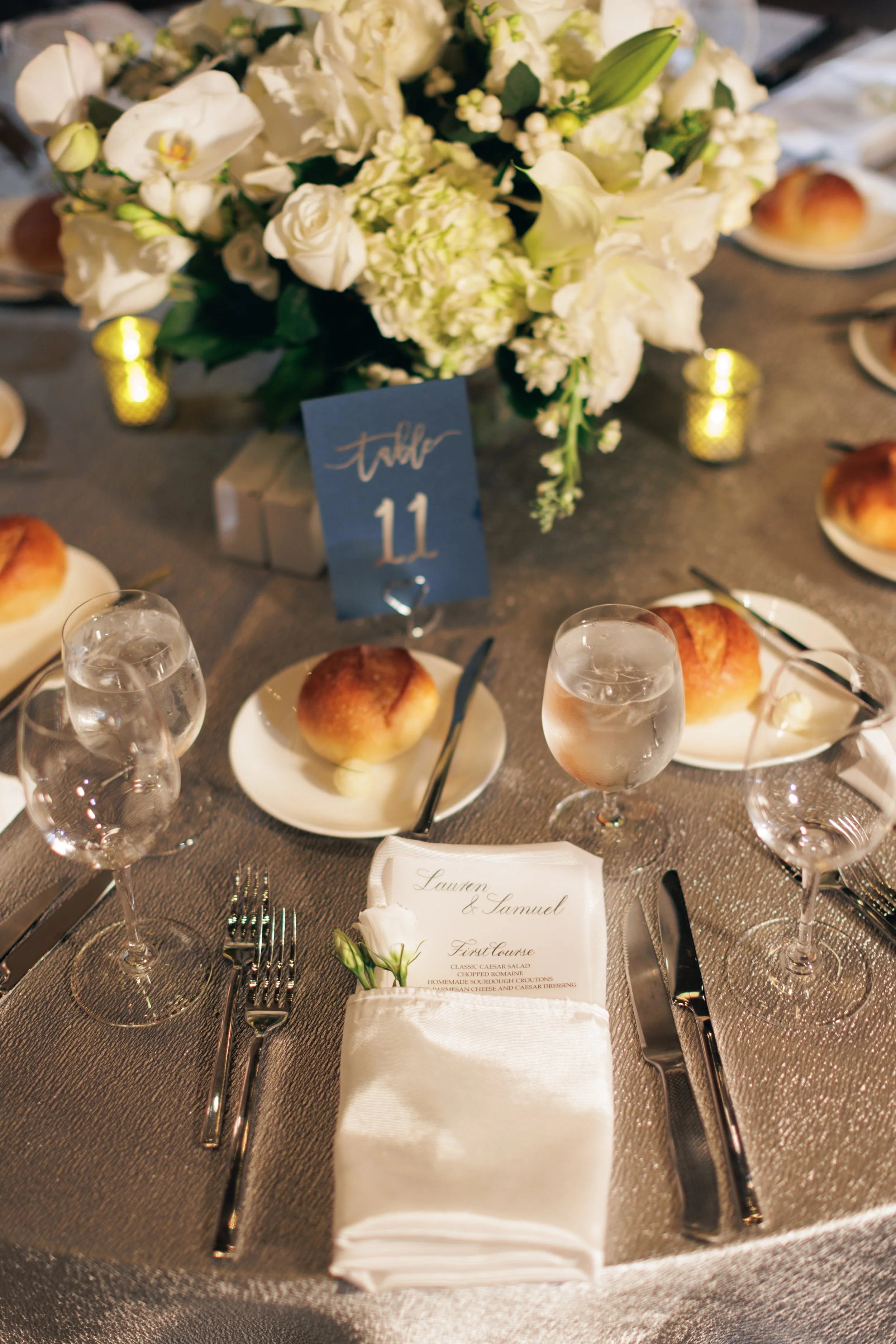 A wedding reception table setting with a floral centerpiece of white roses, lilies, and other flowers, surrounded by candles, glasses of water, silverware, and a folded napkin with a menu. A table number card indicates table 11, and a menu lists dish